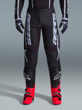 Alpinestars 2026 Fluid Apex Pants, calças de motocross pretas com letras brancas grandes, usadas com proteção de joelho preta e botas vermelhas vibrantes, chassis leve em polifabric 600D, design sport-fit para corridas todo-o-terreno