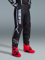 Calças Alpinestars 2026 Fluid Apex, calças de motocross, pretas e cinzentas, usadas com botas rosa vibrante e vermelhas e camisola preta e branca a combinar, com a marca Alpinestars vertical e arrojada na coxa, painéis reforçados e fecho de catraca na cintura para desempenho todo-o-terreno.