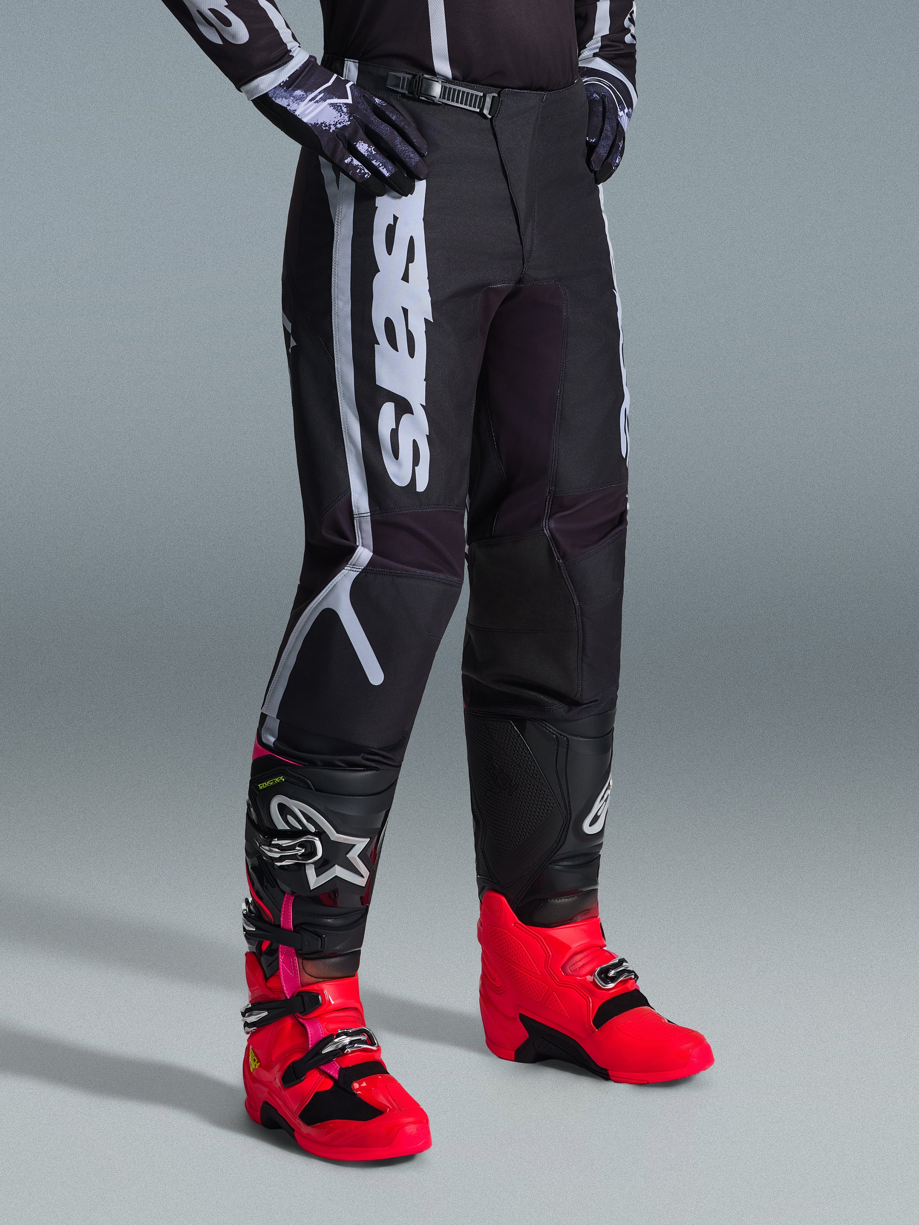 Calças Alpinestars 2026 Fluid Apex, calças de motocross, pretas e cinzentas, usadas com botas rosa vibrante e vermelhas e camisola preta e branca a combinar, com a marca Alpinestars vertical e arrojada na coxa, painéis reforçados e fecho de catraca na cintura para desempenho todo-o-terreno.
