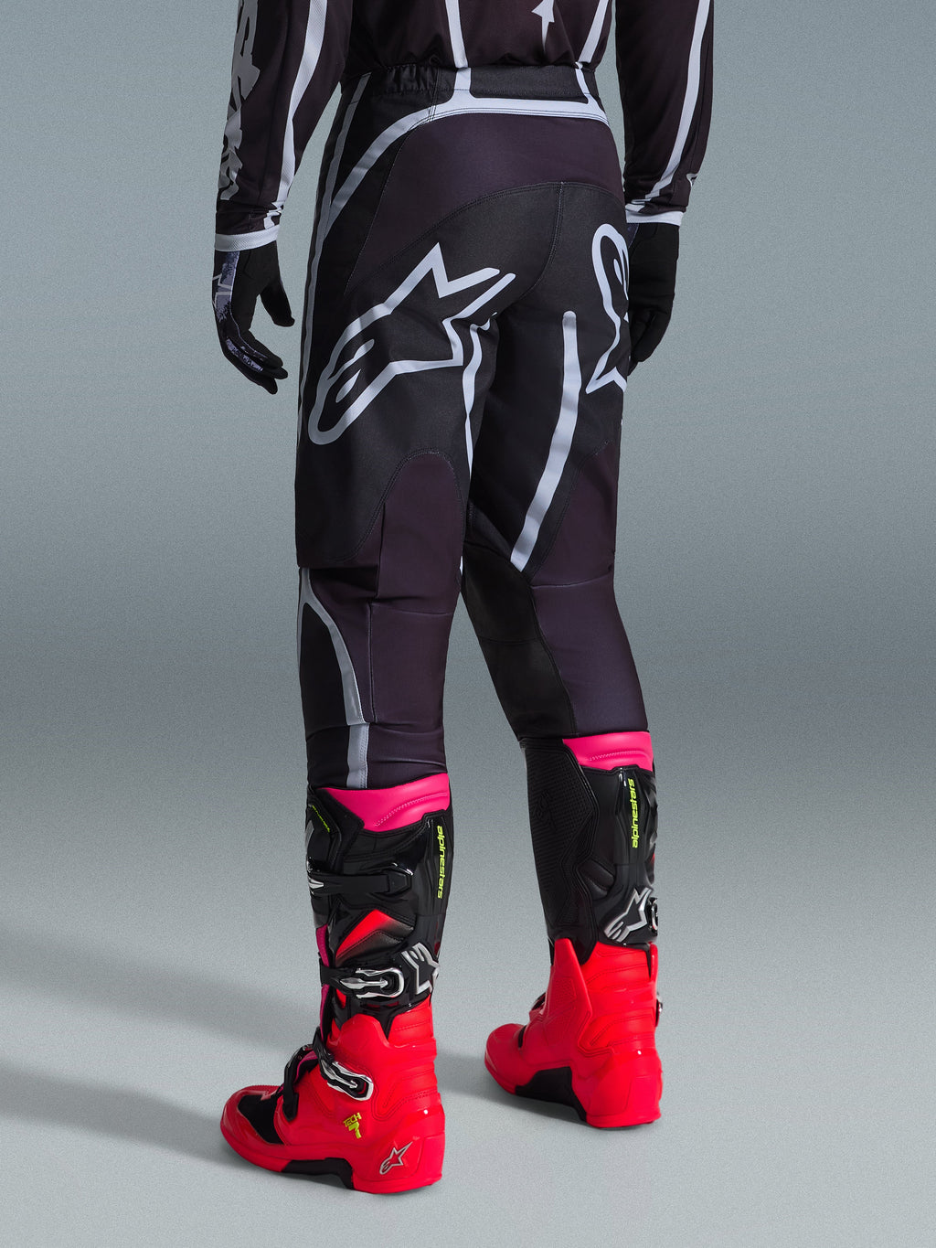 Alpinestars 2026 Fluid Apex Pants, calças de motocross, pretas e cinzentas, vista traseira com grandes logótipos Alpinestars brancos no assento, usadas com botas vermelhas e cor-de-rosa vibrantes, construção em polifabric 600D durável para desempenho em todo-o-terreno