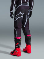 Alpinestars 2026 Fluid Apex Pants, calças de motocross, pretas e cinzentas, vista traseira com grandes logótipos Alpinestars brancos no assento, usadas com botas vermelhas e cor-de-rosa vibrantes, construção em polifabric 600D durável para desempenho em todo-o-terreno