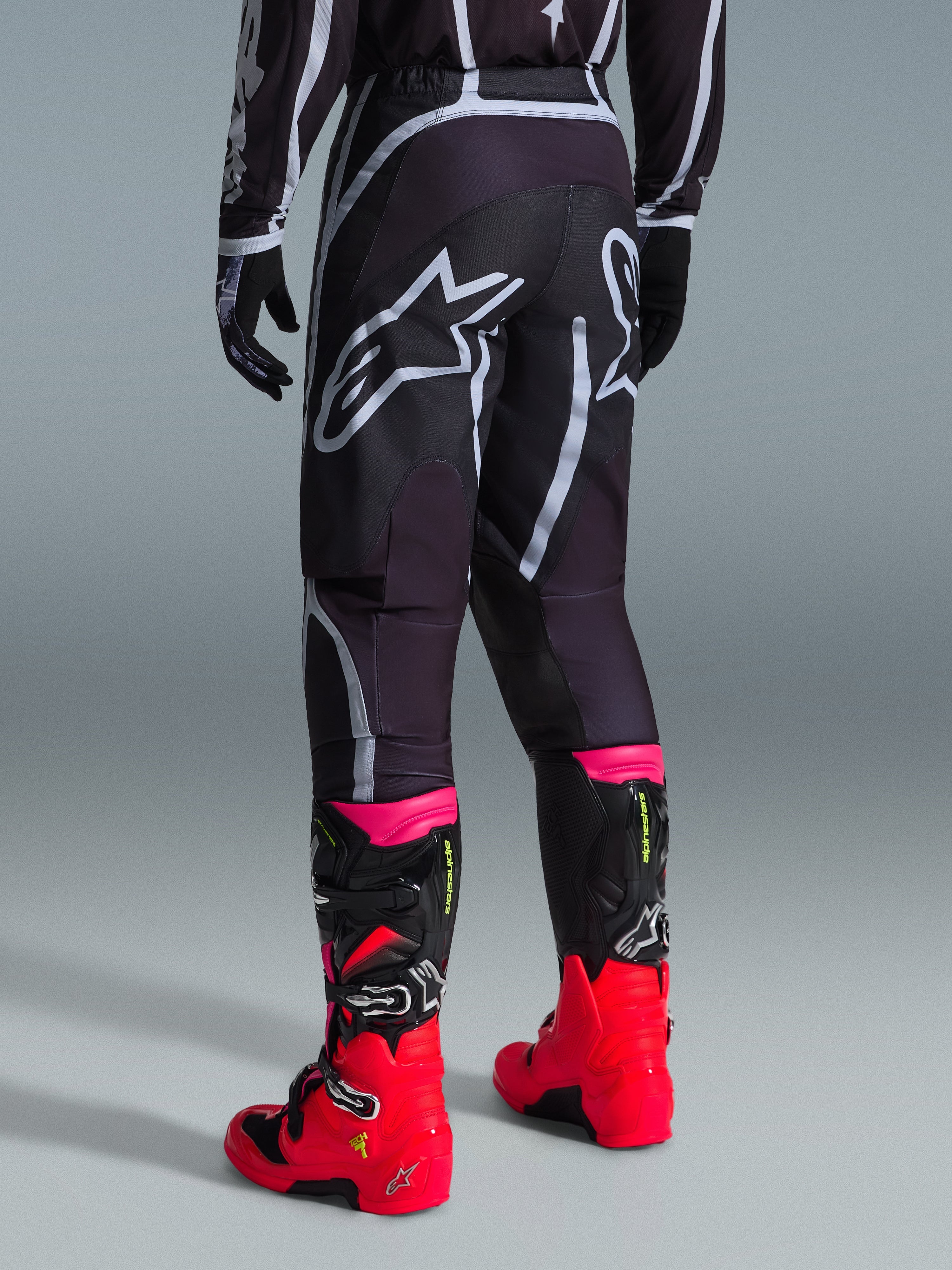 Alpinestars 2026 Fluid Apex Pants, calças de motocross, pretas e cinzentas, vista traseira com grandes logótipos Alpinestars brancos no assento, usadas com botas vermelhas e cor-de-rosa vibrantes, construção em polifabric 600D durável para desempenho em todo-o-terreno