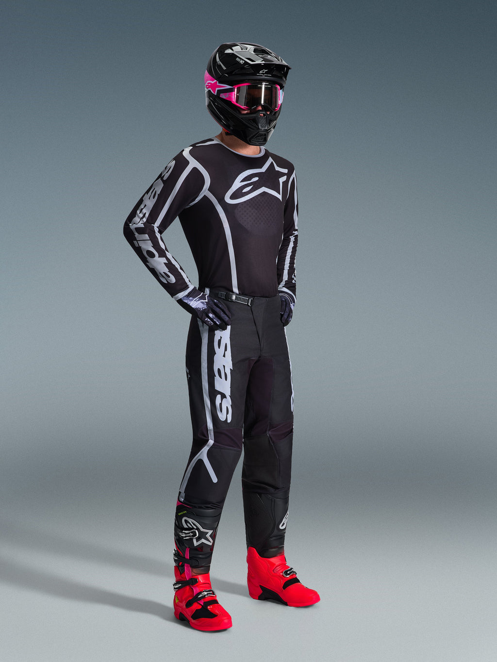 Alpinestars 2026 Fluid Apex Pants, calças de motocross, pretas e cinzentas, usadas com camisola preta e branca a condizer, capacete preto com óculos cor-de-rosa e botas vermelhas vibrantes, com logótipos brancos em destaque nas pernas e design de ajuste desportivo para corridas off-road