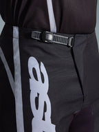 Calças Alpinestars 2026 Fluid Apex, calças de motocross, pretas e cinzentas, pormenor aproximado do sistema de fecho de catraca integrado para um ajuste seguro, com grafismos do logótipo brancos em polifabric 600D durável.