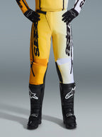 Calças Alpinestars 2026 Fluid Apex, calças de motocross, amarelas e pretas, design de cores divididas com painéis amarelos e brancos, marca vertical 'astars' em destaque, usadas com botas e luvas de motocross pretas, equipamento de performance para condução todo-o-terreno