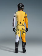 Calças Alpinestars 2026 Fluid Apex, calças de motocross, amarelas e pretas, vista traseira de um piloto com equipamento completo de motocross, camisola a condizer com gradientes em branco e laranja, capacete preto, botas pretas e luvas pretas, conjunto de todo-o-terreno coordenado.