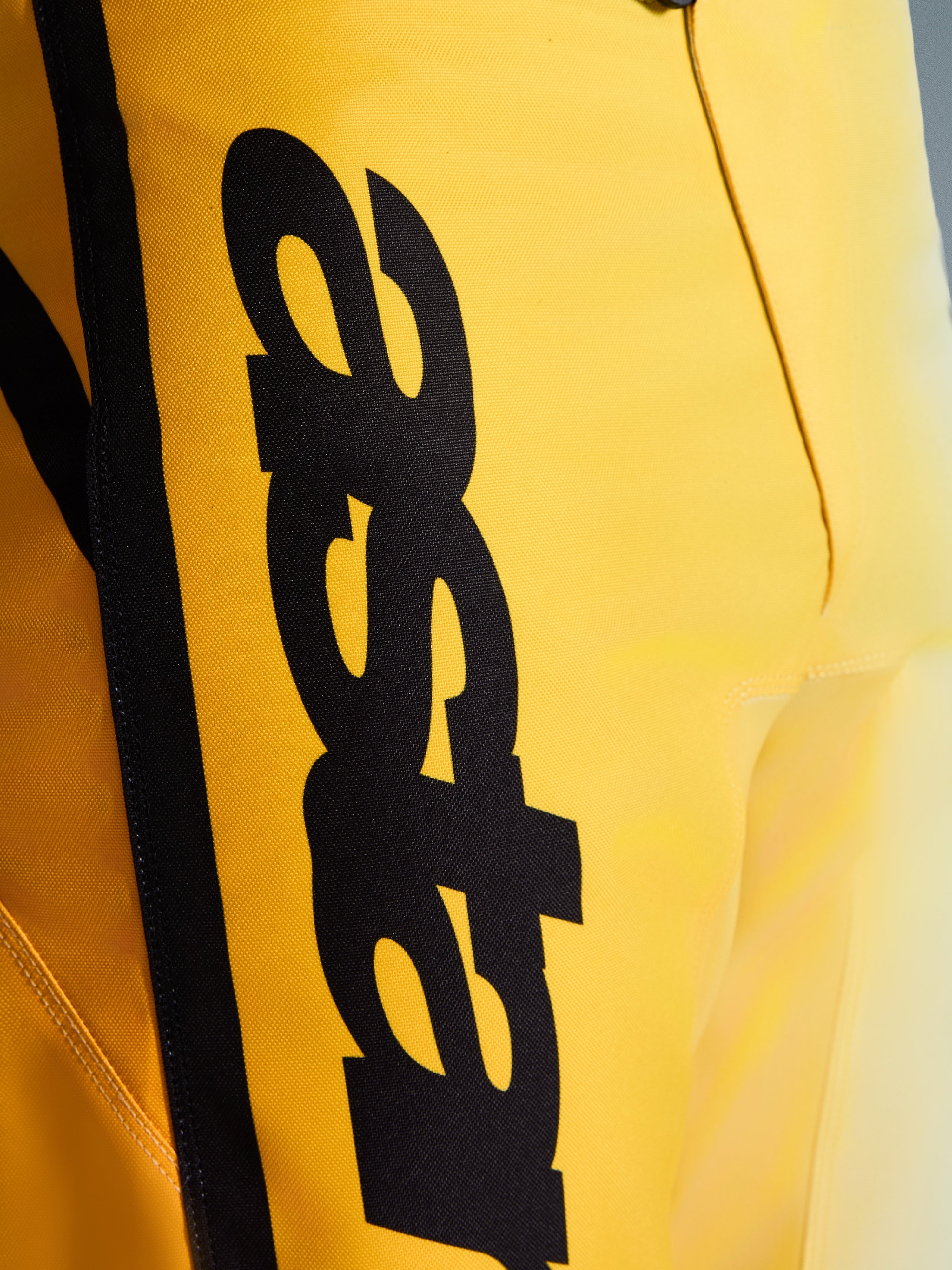 Close-up das calças Alpinestars 2026 Fluid Apex, calças de motocross, amarelas e pretas, com um grande logótipo preto vertical, material durável texturizado e detalhes ergonómicos para desempenho todo-o-terreno