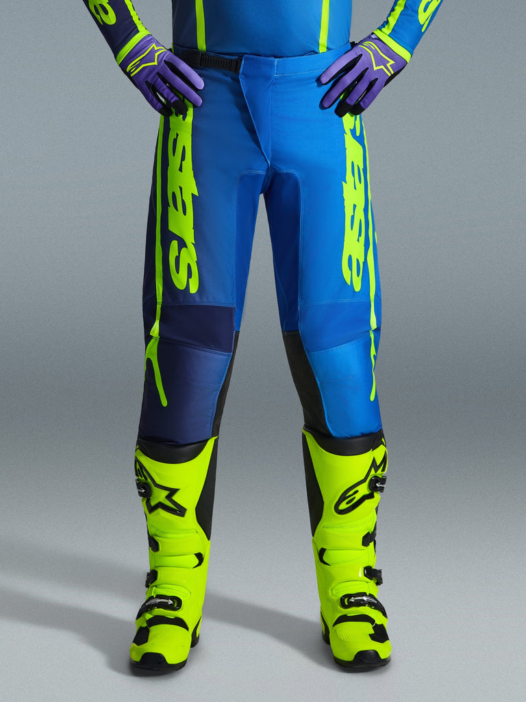 Alpinestars 2026 Fluid Apex Pants, calças de motocross, azul e amarelo fluo, usadas com botas amarelas fluo a condizer e luvas roxas, logótipo amarelo de destaque nas laterais, design ergonómico para corridas todo-o-terreno