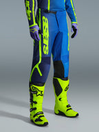 Alpinestars 2026 Fluid Apex Pants, calças de motocross, azul e amarelo fluo, usadas com luvas a condizer e botas amarelo fluo brilhante, com logótipo vertical de destaque, painéis de joelho reforçados e fecho de cintura de catraca seguro