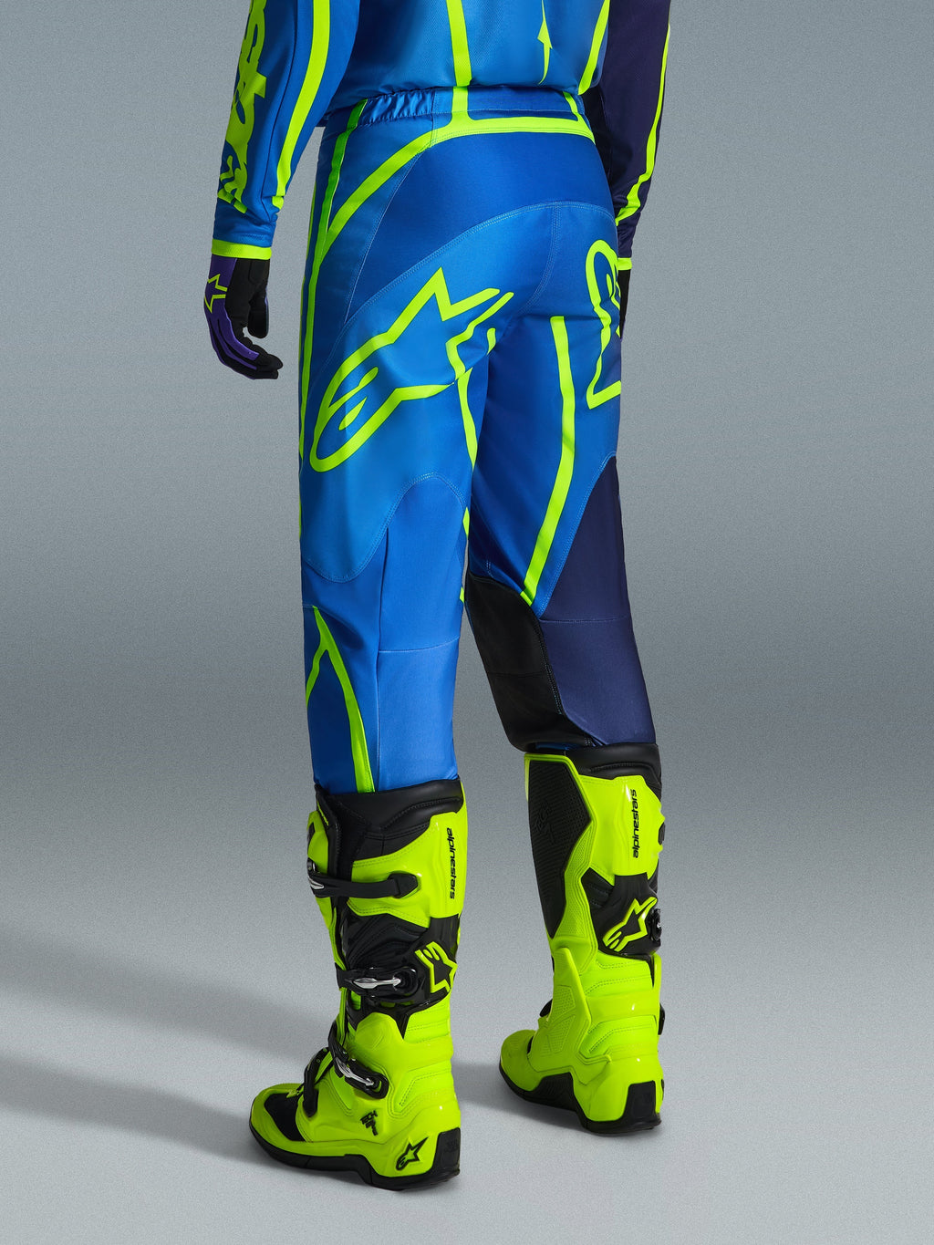 Alpinestars 2026 Fluid Apex Pants, calças de motocross, azul e amarelo néon, vista traseira do piloto a usar equipamento a condizer e botas amarelo fluorescente, com logótipos Alpinestars marcantes e design ergonómico