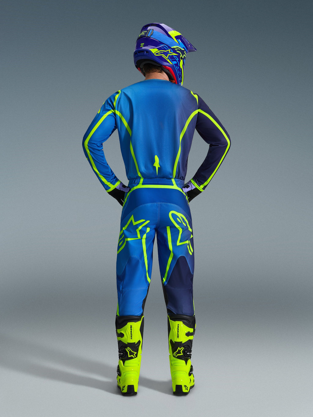 Alpinestars 2026 Fluid Apex Pants, calças de motocross, azuis e amarelo fluo, vista traseira de um piloto com equipamento de corrida completo, camisola e capacete a combinar, botas amarelo brilhante, corte atlético coordenado para motocross