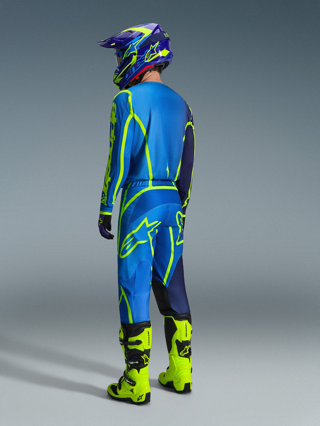 Piloto de motocross visto de costas a usar calças Alpinestars Fluid Apex 2026 em azul e amarelo fluo, camisola e capacete a condizer em azul e amarelo, e botas em amarelo néon brilhante, equipamento de corrida coordenado para desempenho em todo-o-terreno