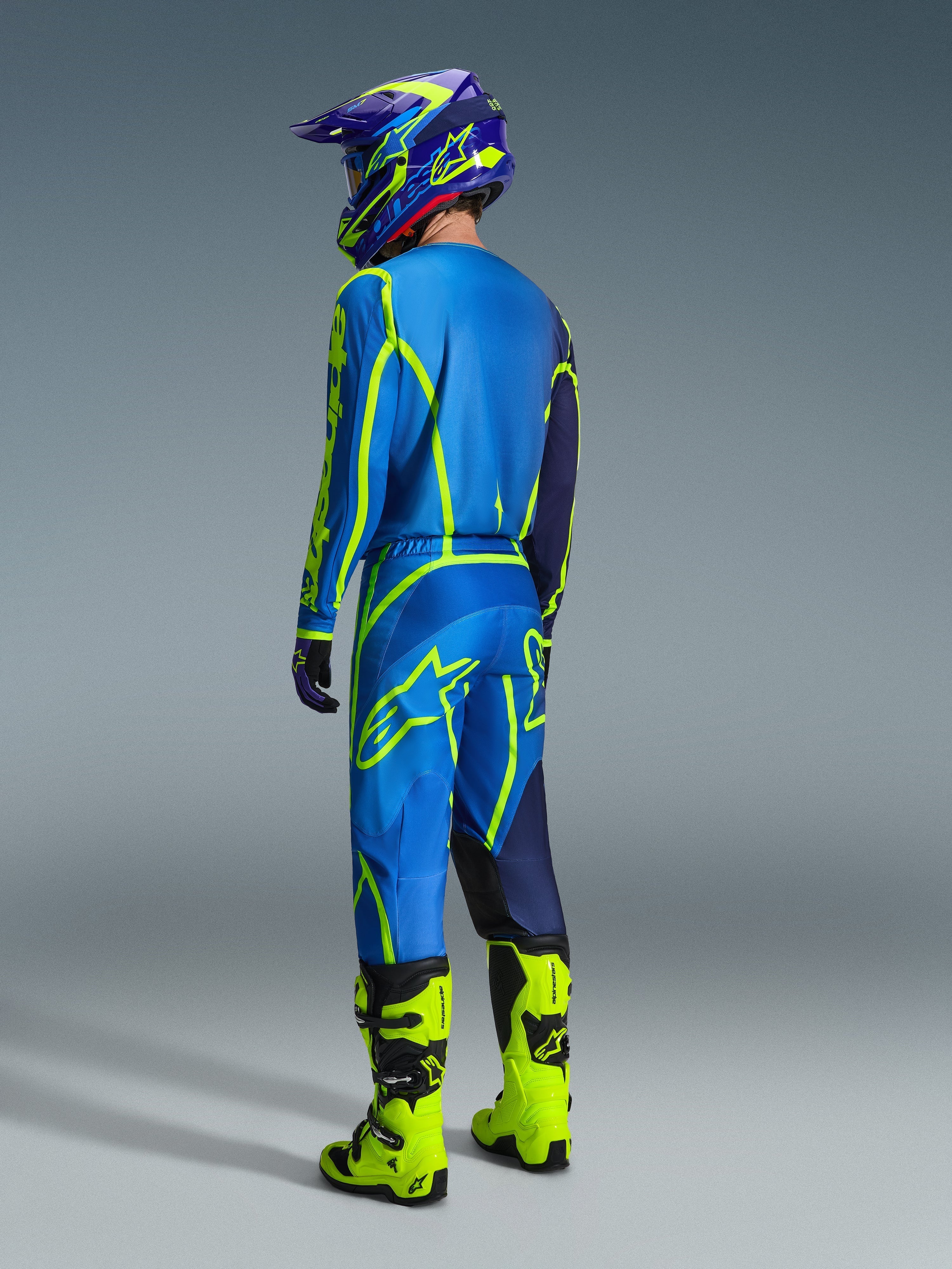 Piloto de motocross visto de costas a usar calças Alpinestars Fluid Apex 2026 em azul e amarelo fluo, camisola e capacete a condizer em azul e amarelo, e botas em amarelo néon brilhante, equipamento de corrida coordenado para desempenho em todo-o-terreno