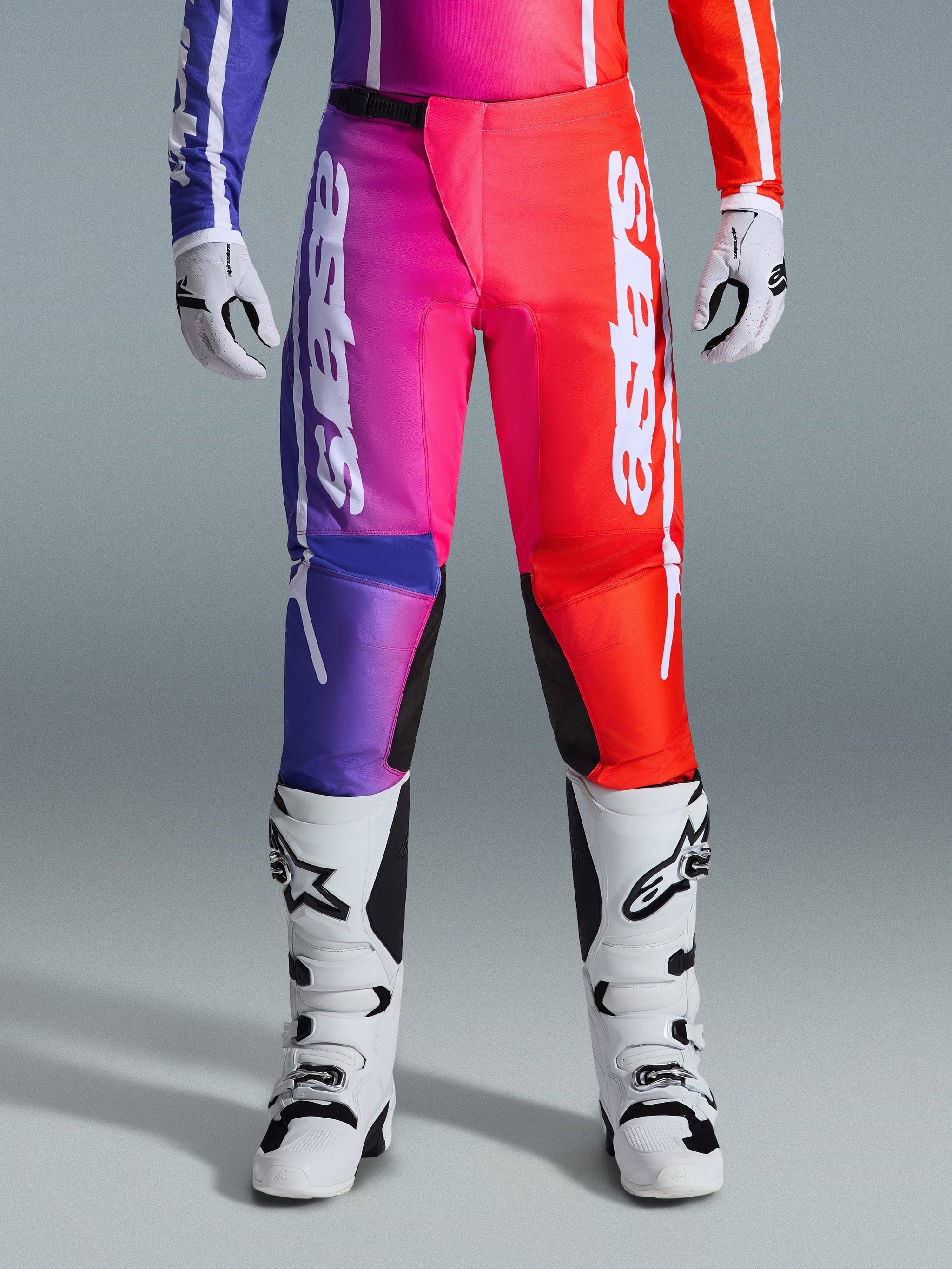 Alpinestars 2026 Fluid Apex Pants, calças de motocross, design degradê multicolorido em roxo, rosa e vermelho, logótipo Alpinestars branco de grandes dimensões nas pernas, combinadas com botas e luvas de motocross brancas e pretas, equipamento profissional de corrida todo-o-terreno