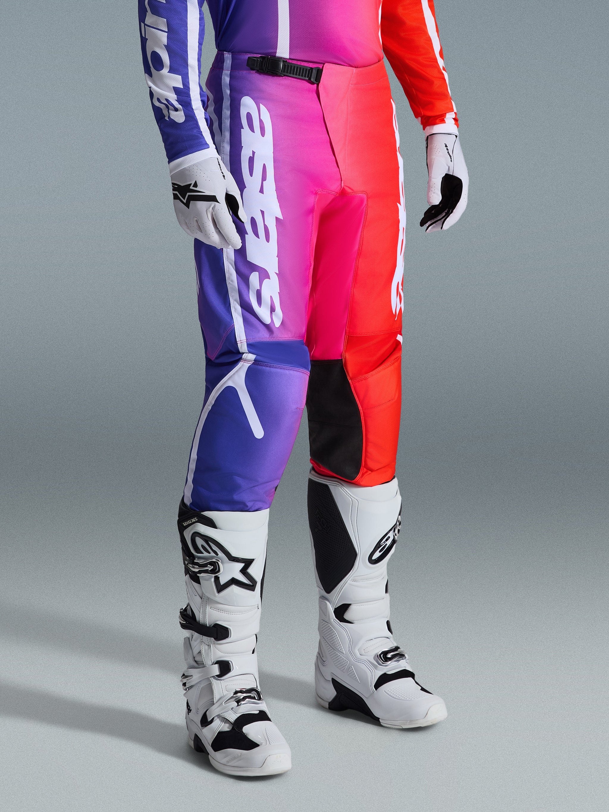 Alpinestars 2026 Fluid Apex Pants, calças de motocross, gradiente multicolorido em roxo, rosa e vermelho, usadas com botas e luvas de motocross brancas e pretas, com marca vertical branca e detalhes pretos nos joelhos, equipamento de performance para todo-o-terreno