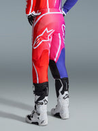 Alpinestars 2026 Fluid Apex Pants, calças de motocross, multicolor em vermelho, rosa e roxo, vista traseira de piloto com botas de motocross brancas e pretas, com logótipo Alpinestars branco de grandes dimensões e design de gradiente vibrante