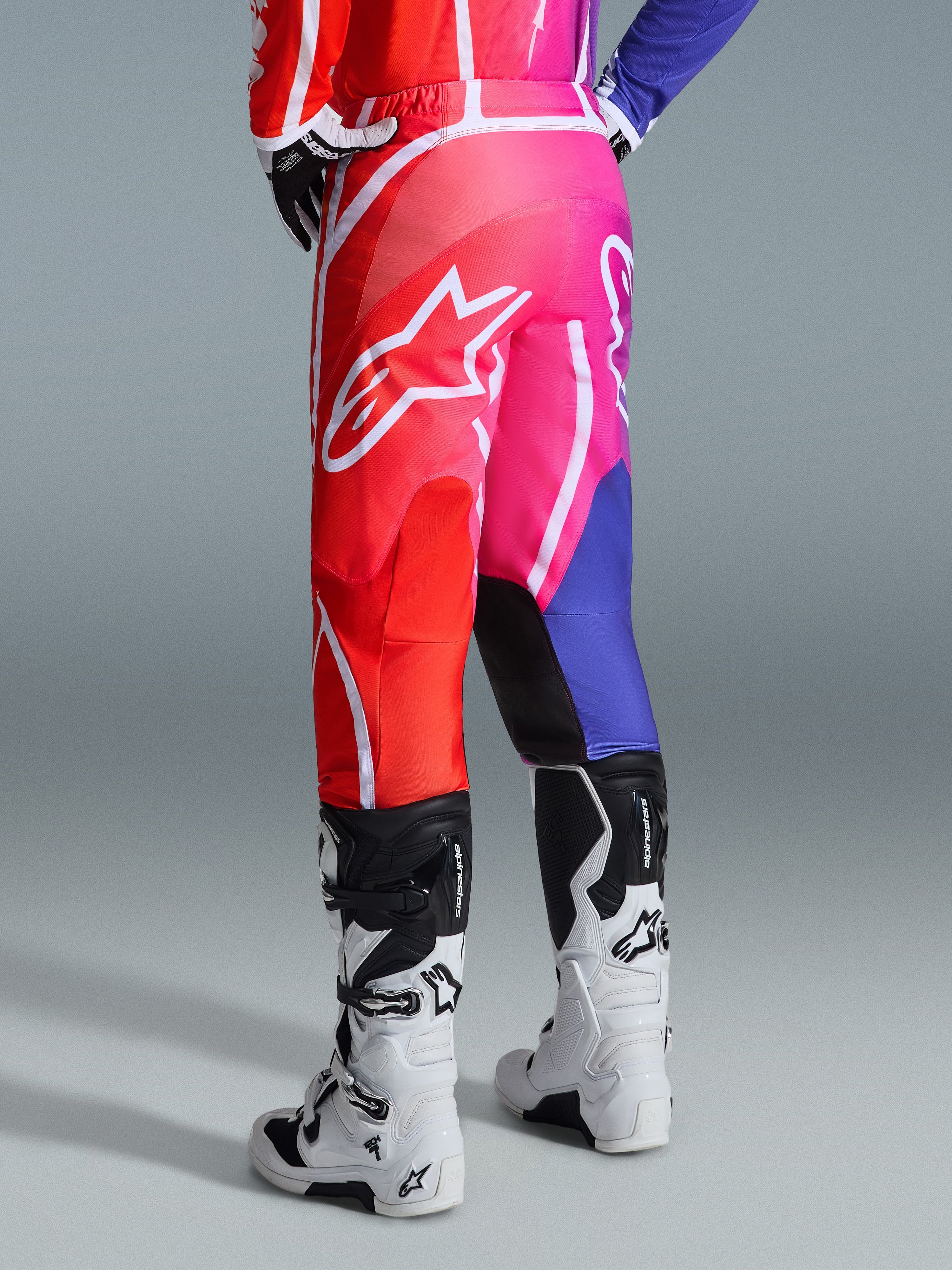 Alpinestars 2026 Fluid Apex Pants, calças de motocross, multicolor em vermelho, rosa e roxo, vista traseira de piloto com botas de motocross brancas e pretas, com logótipo Alpinestars branco de grandes dimensões e design de gradiente vibrante