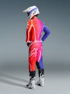 Piloto de motocross com equipamento Alpinestars 2026 Fluid Apex, camisola e calças multicolor com gradiente de vermelho, rosa e roxo, logótipos brancos, capacete branco e botas brancas, conjunto coordenado para corridas fora de estrada