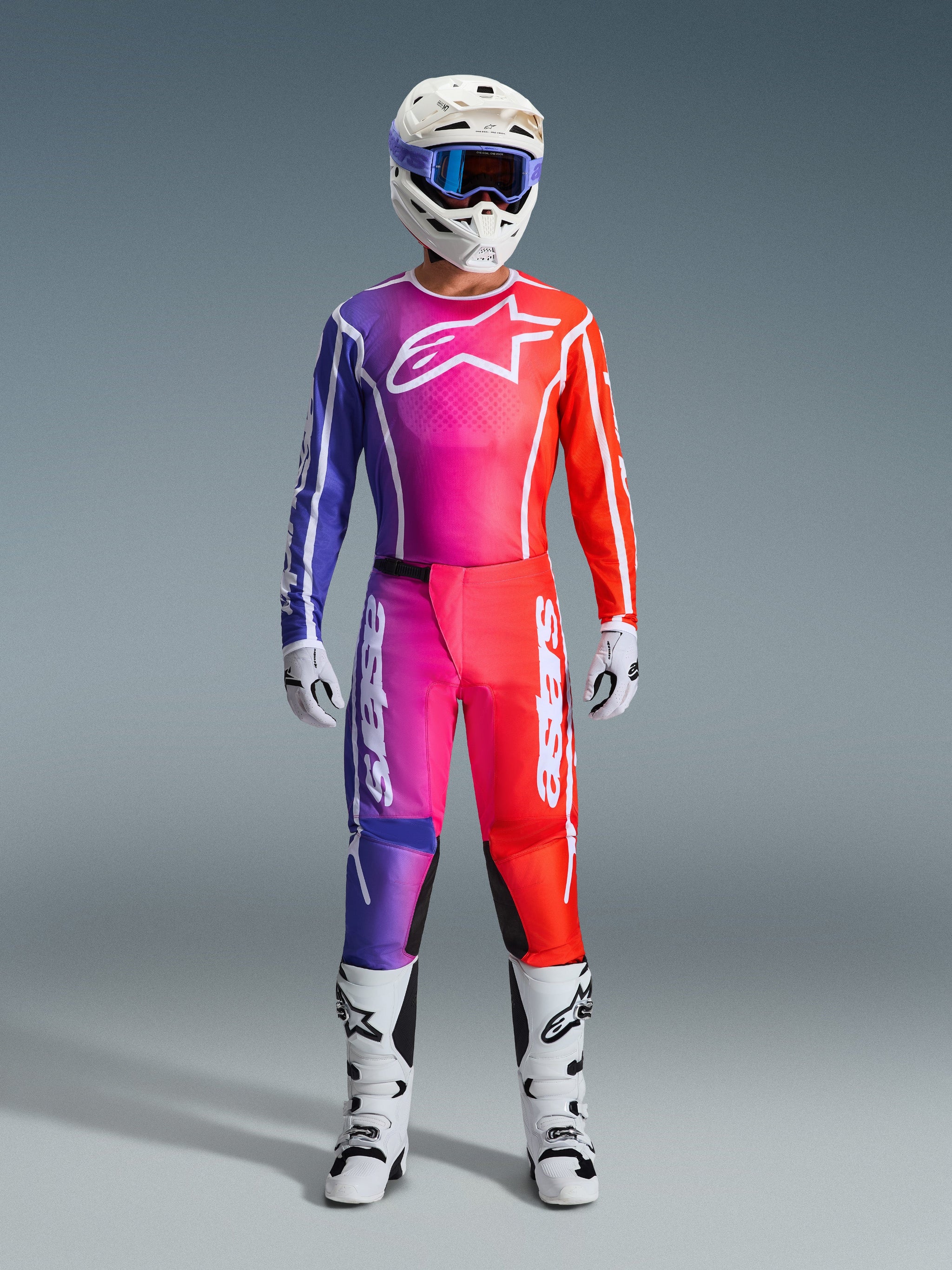Piloto de motocross com calças Fluid Apex 2026 e camisola a condizer, degradé de roxo e laranja, capacete branco com óculos, botas e luvas brancas, equipamento de corrida Alpinestars coordenado para desempenho em todo-o-terreno.