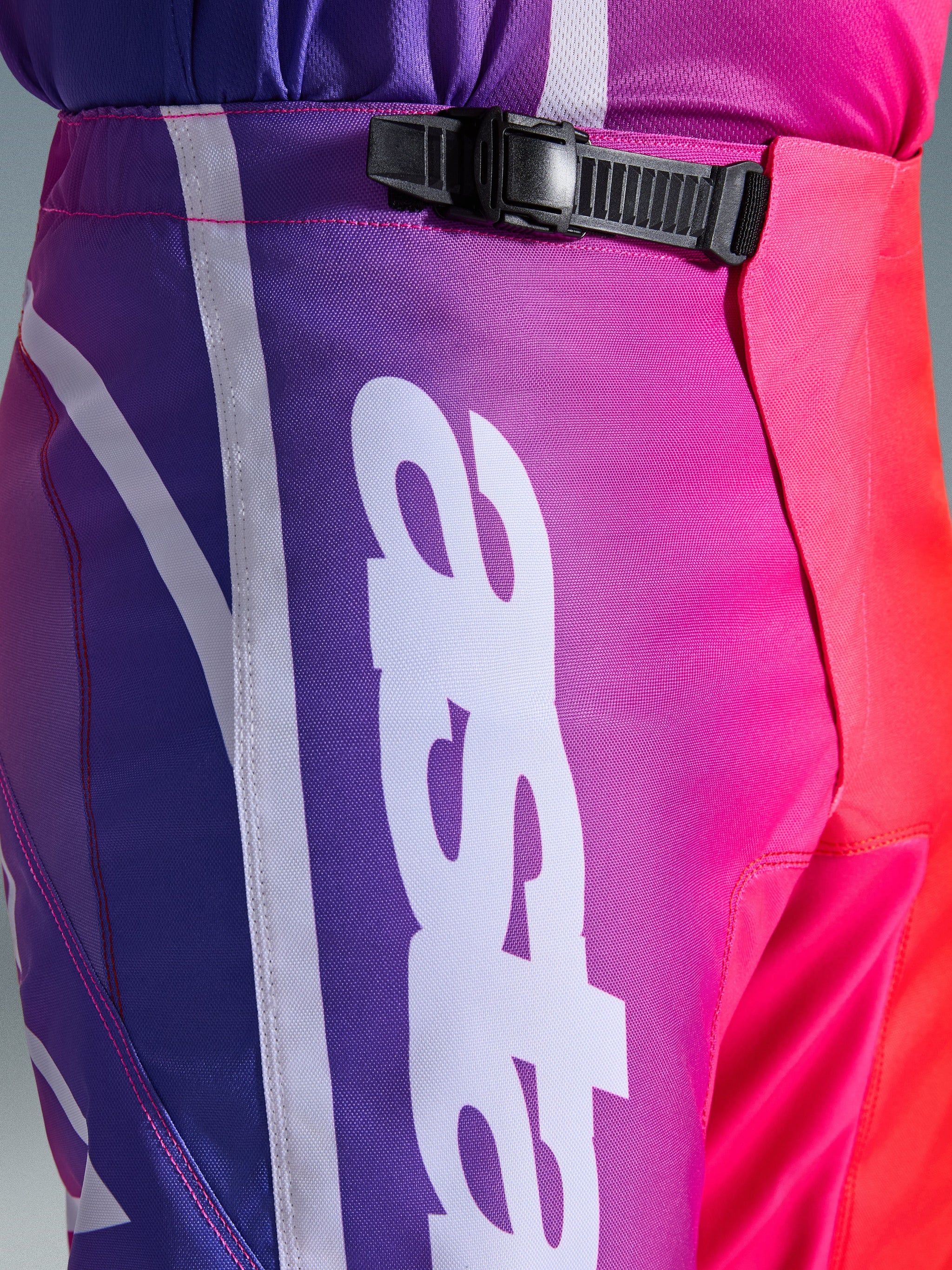 Alpinestars 2026 Fluid Apex Pants, calças de motocross, multicoloridas em roxo, cor-de-rosa e vermelho, grande plano da zona da cintura e anca com um sistema de fecho de catraca preto e grande logótipo Alpinestars vertical em branco, design com gradiente vibrante e textura durável em polifabric