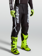 Calças Alpinestars Fluid Apex V2, equipamento de motocross para homem, pretas e prateadas, com botas amarelas vibrantes.