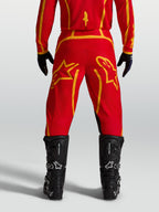 Alpinestars Calças Fluid Apex V2, calças de motocross para homem, vermelhas e douradas, vista traseira com botas de moto pretas.