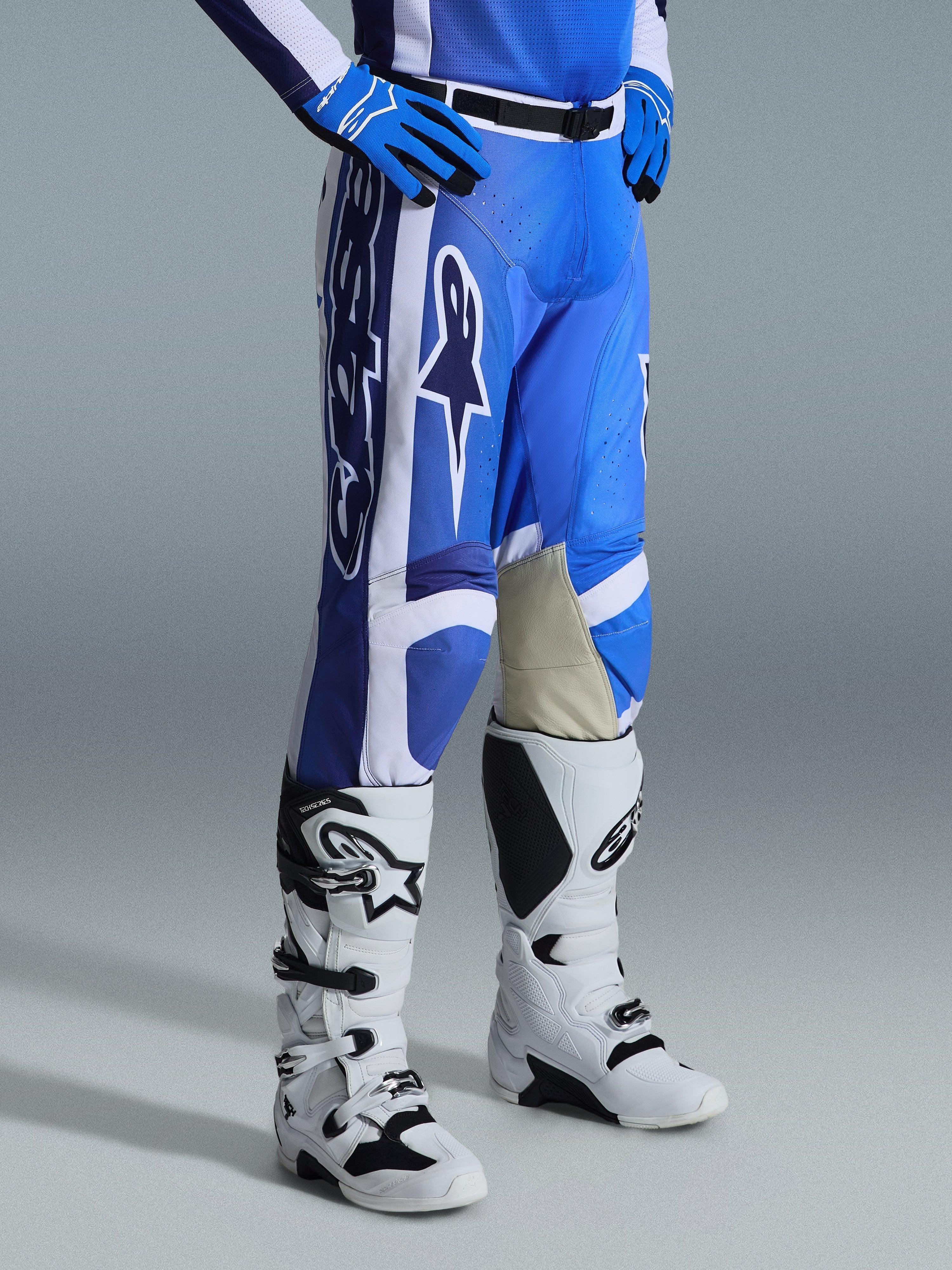 Calças Alpinestars 2026 Racer Air Portl, calças de motocross, azul Ucla e branco, usadas com luvas azuis a condizer e botas de motocross brancas e pretas, com logótipos Alpinestars brancos de grandes dimensões, painéis interiores dos joelhos em pele bege e uma fivela magnética preta na cintura.