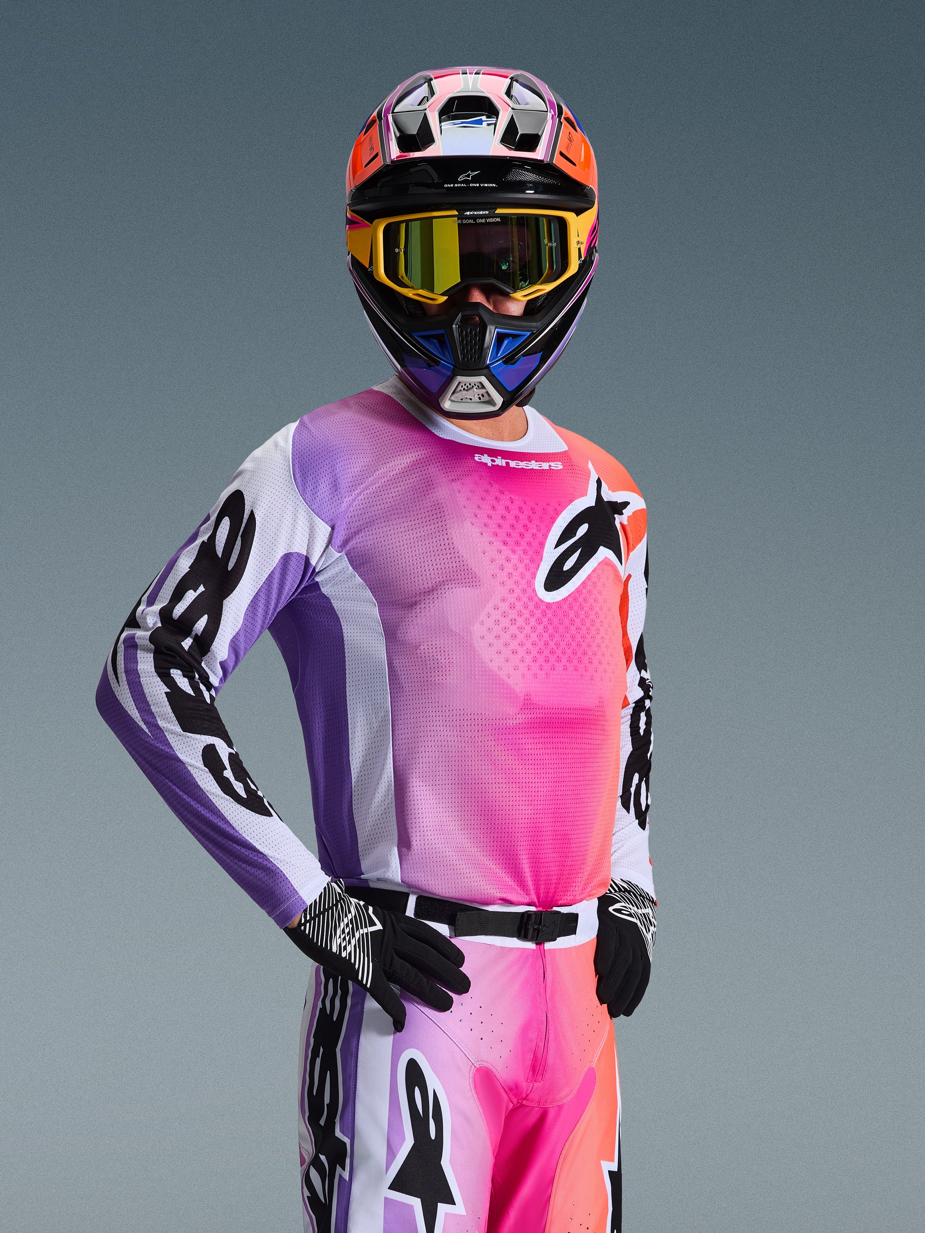 Piloto de motocross com a camisola Alpinestars 2026 Racer Air Portl, degradê multicolorido em rosa, roxo e laranja com detalhes brancos, capacete colorido a condizer, óculos amarelos, luvas pretas e equipamento de corrida off-road profissional