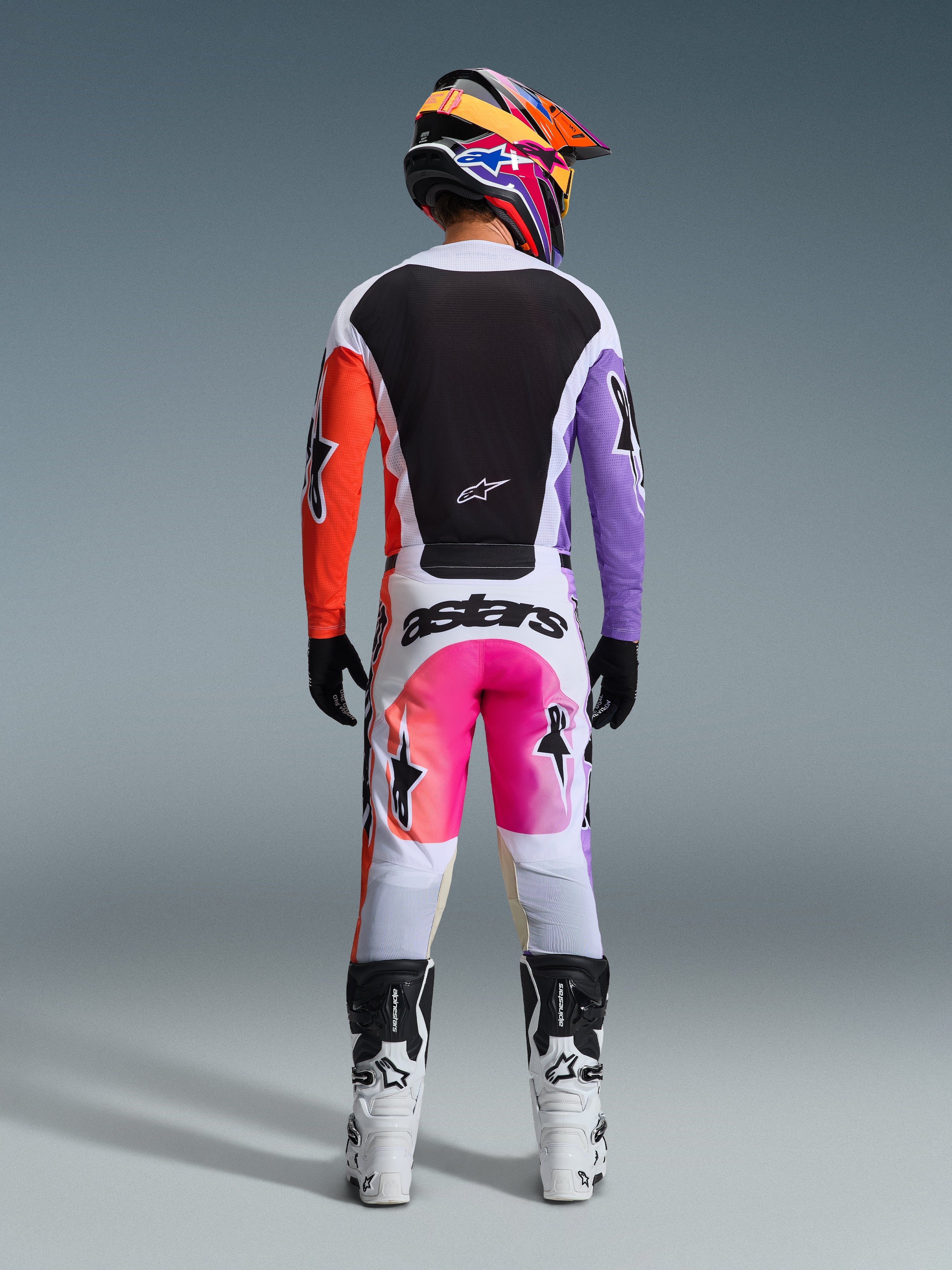 Piloto de motocross com equipamento completo de corrida visto de trás, camisola Alpinestars 2026 Racer Air Portl com painéis brancos, pretos, cor de laranja e roxos, calças brancas a condizer com detalhes cor-de-rosa e marca 'astars', botas brancas e pretas e um capacete multicolorido vibrante, conjunto coordenado para corridas de motocross