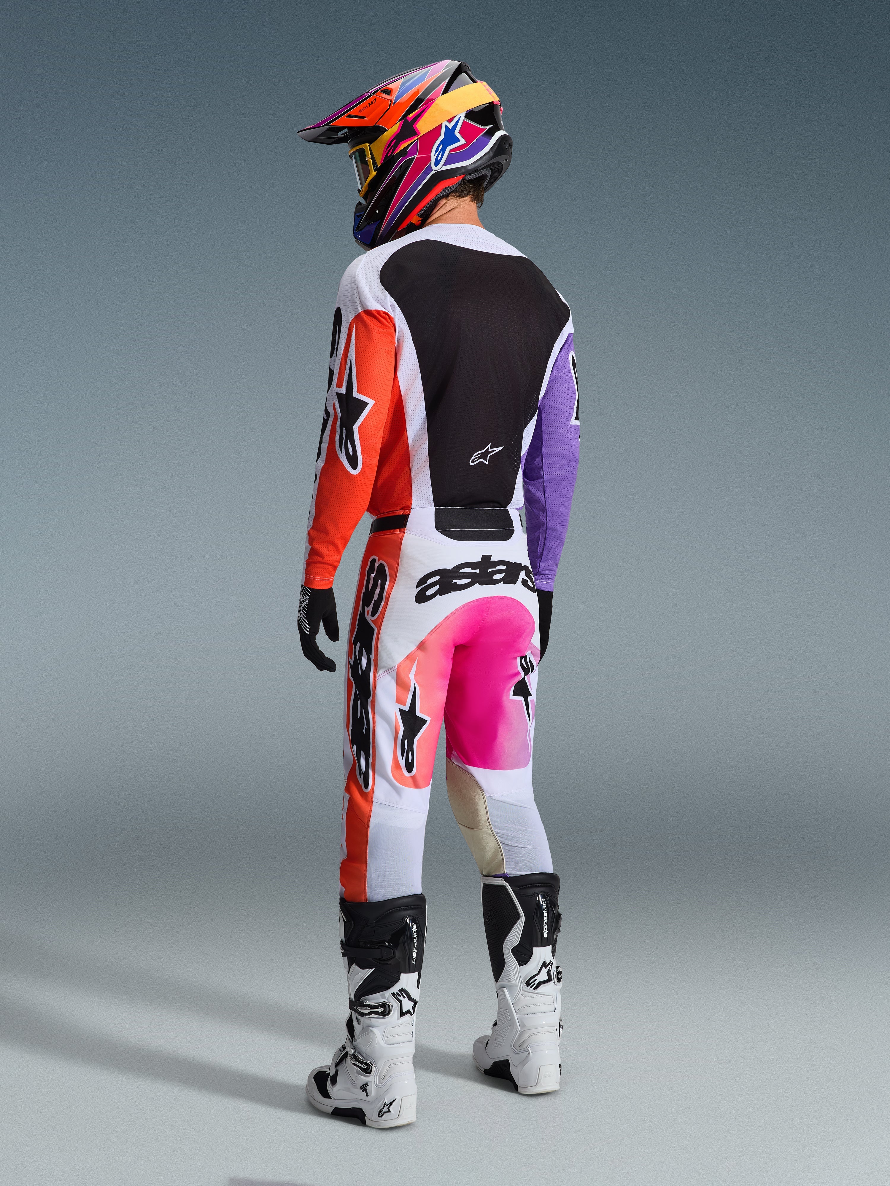 Alpinestars 2026 Racer Air Portl Jersey, camisola de motocross multicolor, vista traseira de um piloto com equipamento de corrida completo, apresentando base branca com mangas cor de laranja e roxas e painel traseiro em malha preta, combinada com calças cor-de-rosa e laranja a condizer, capacete colorido e botas brancas, conjunto de corrida profissional de off-road