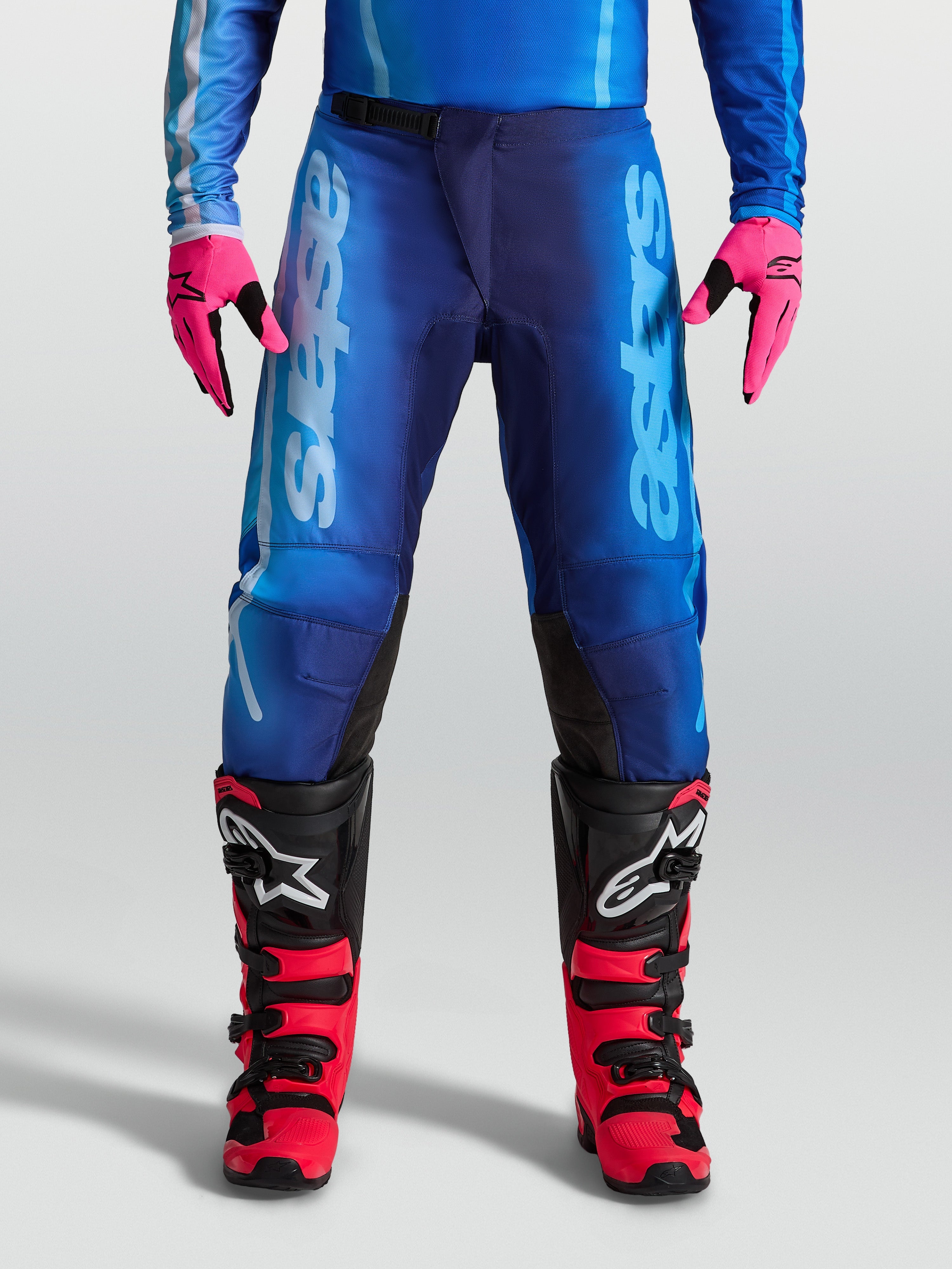 Alpinestars Calças Fluid Kepler, calças de motocross para homem, azuis, em polifabric 600D leve com ajuste ergonómico, modelo equipado com luvas cor-de-rosa e botas de mota vermelhas e pretas.