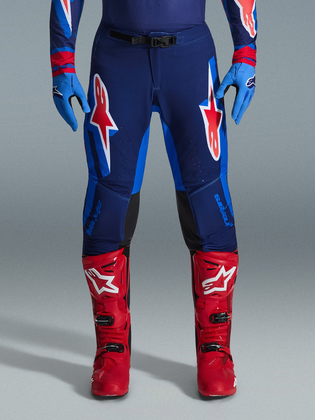 Alpinestars 2026 Supertech Vista Pants, calças de motocross, em azul-escuro, coral vivo e cobalto, vista frontal de piloto com equipamento de alta performance, luvas azuis e botas de motocross vermelhas, com logótipos Alpinestars proeminentes em vermelho e branco nas coxas
