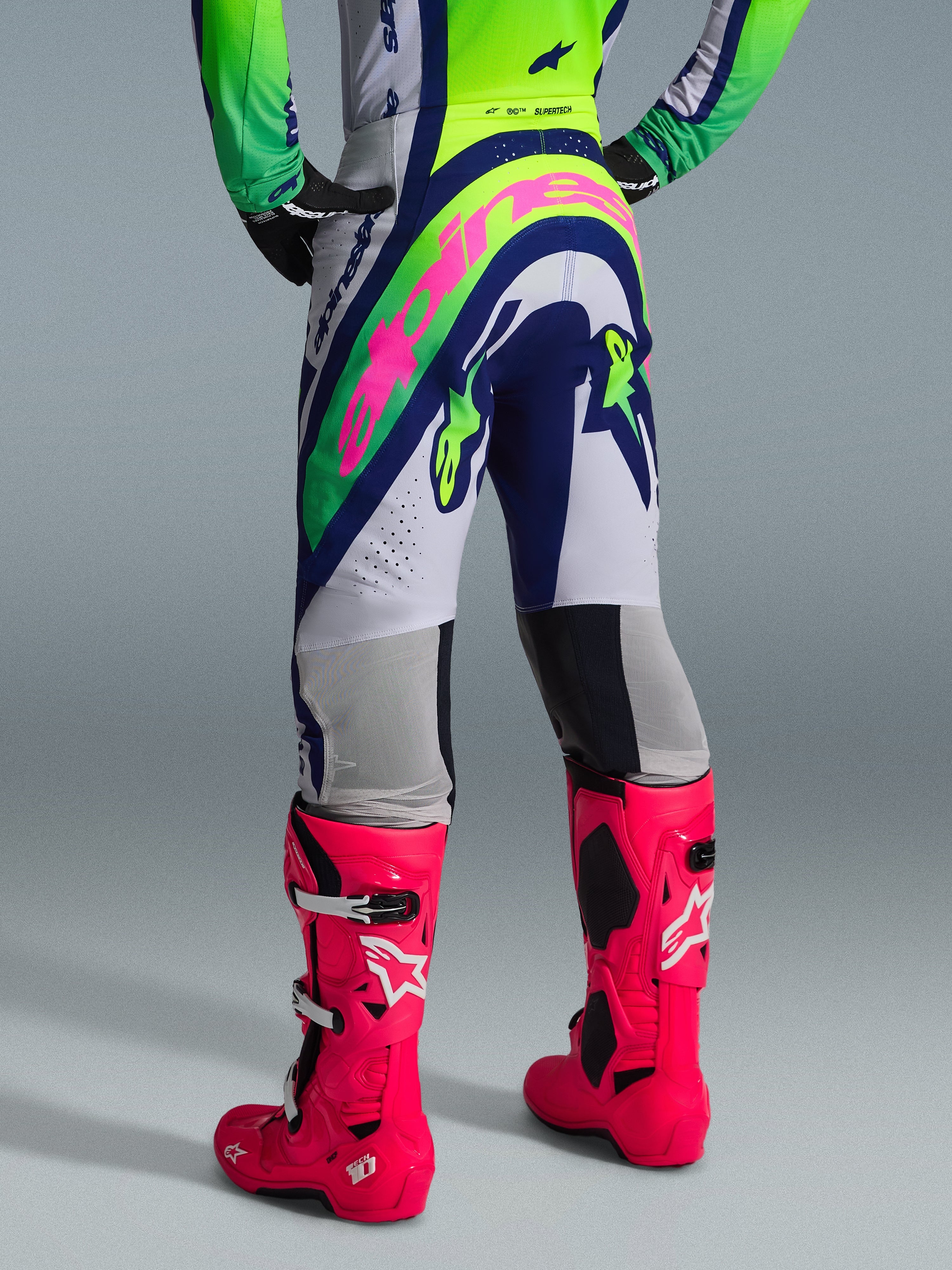 Conjunto de Equipamento Alpinestars 2026 Supertech Vista, vista traseira de vestuário de motocross, calças brancas com painéis azuis e verde-lima, logótipo grande cor-de-rosa na zona do assento, combinado com botas Tech 10 cor-de-rosa vibrante, equipamento profissional de corrida todo-o-terreno