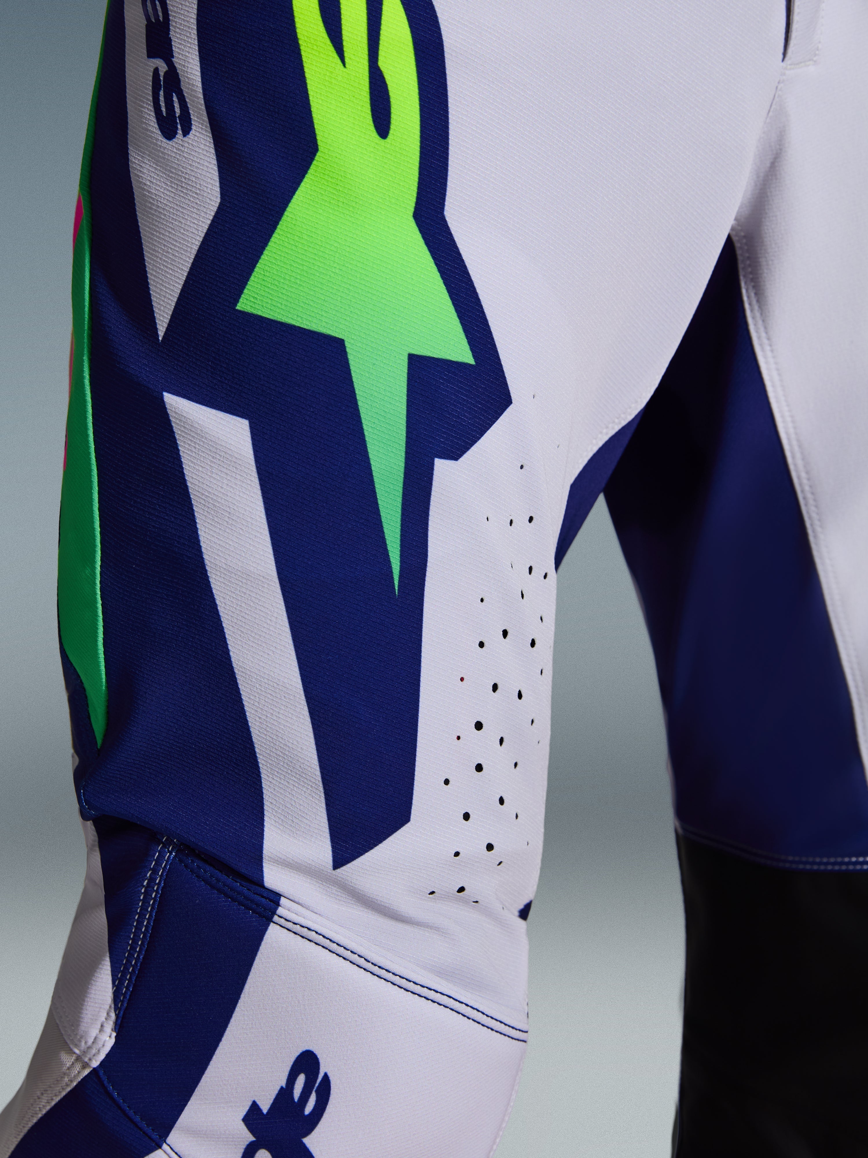 Calças Alpinestars Supertech Vista 2026, calças de motocross, cinzento claro com detalhes em amarelo fluo e azul, vista aproximada da coxa mostrando o logótipo A-star de grandes dimensões, orifícios de ventilação cortados a laser e tecido rip-stop elástico avançado