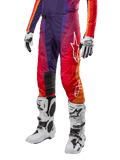 Alpinestars 2024 Techstar Pneuma Pants, calças de motocross em roxo profundo, laranja e azul com gradiente vibrante, logótipo da estrela em branco, joelheiras reforçadas, acompanhadas por botas de motocross brancas e luvas, equipamento de proteção para todo-o-terreno
