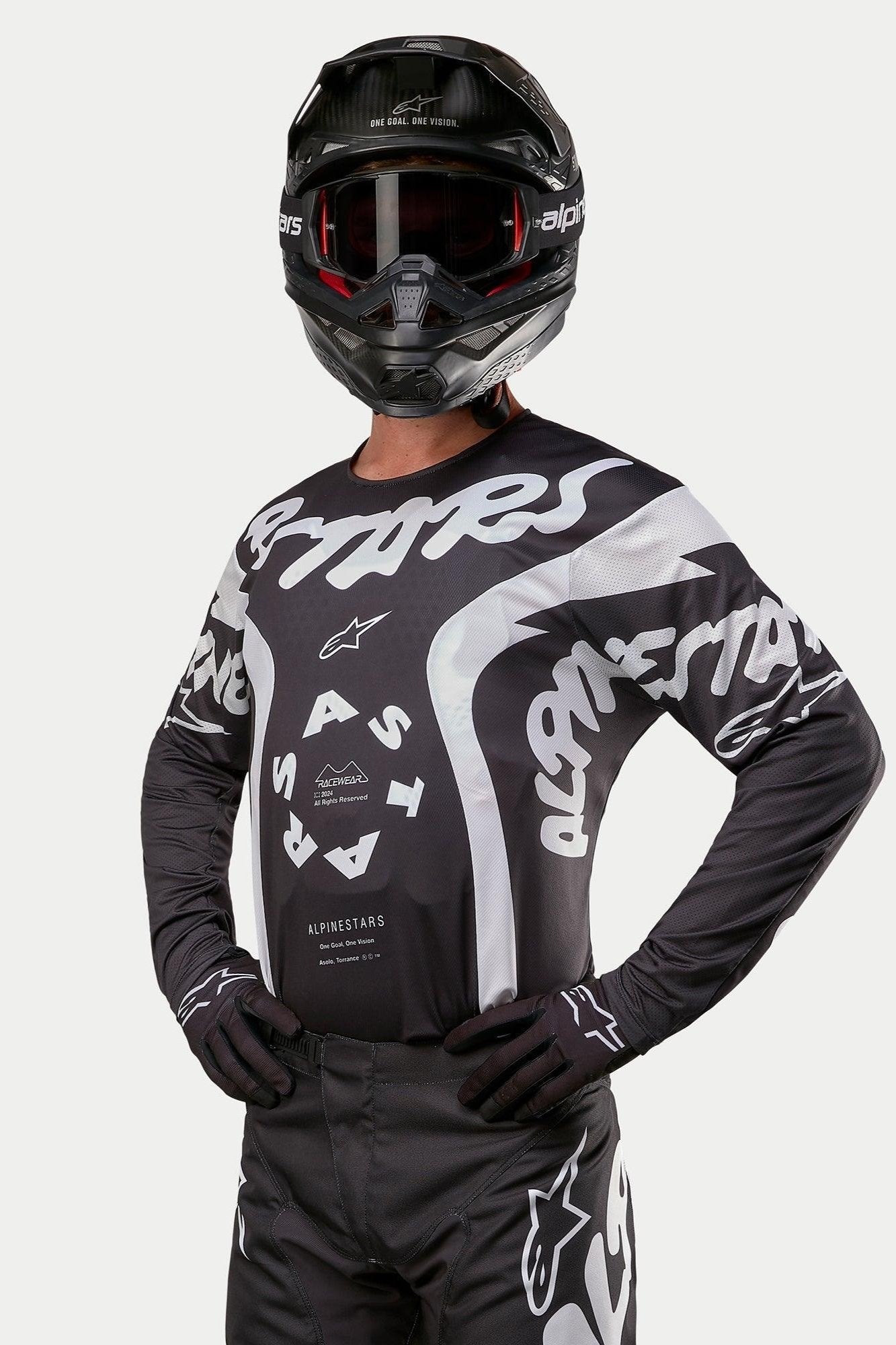 Alpinestars 2024 Racer Hana Jersey, camisola de motocross, preta e branca, com grafismos arrojados de texto e logótipo Alpinestars em branco, usada por um piloto com capacete preto e óculos de proteção, equipamento de alto desempenho para corridas todo-o-terreno