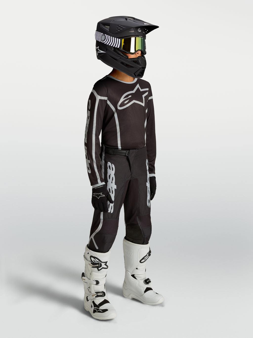 Alpinestars calças Fluid Apex V2 juventude, pretas e prateadas, equipamento de motocross em modelo com botas brancas.