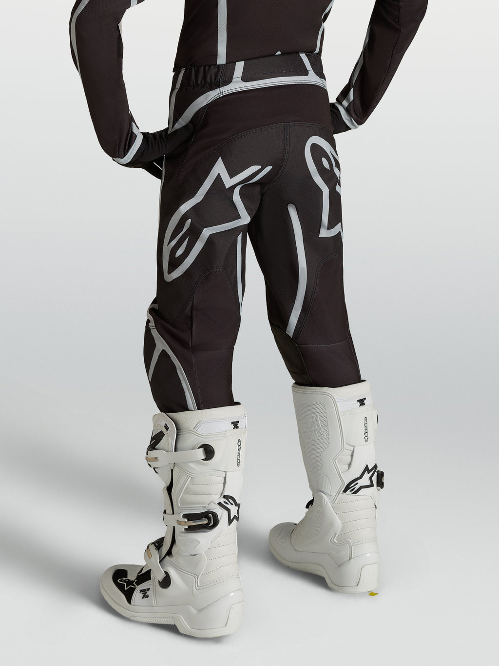 Alpinestars Calças Fluid Apex V2 Juventude, calças de motocross, pretas e prateadas, vista traseira com botas brancas.