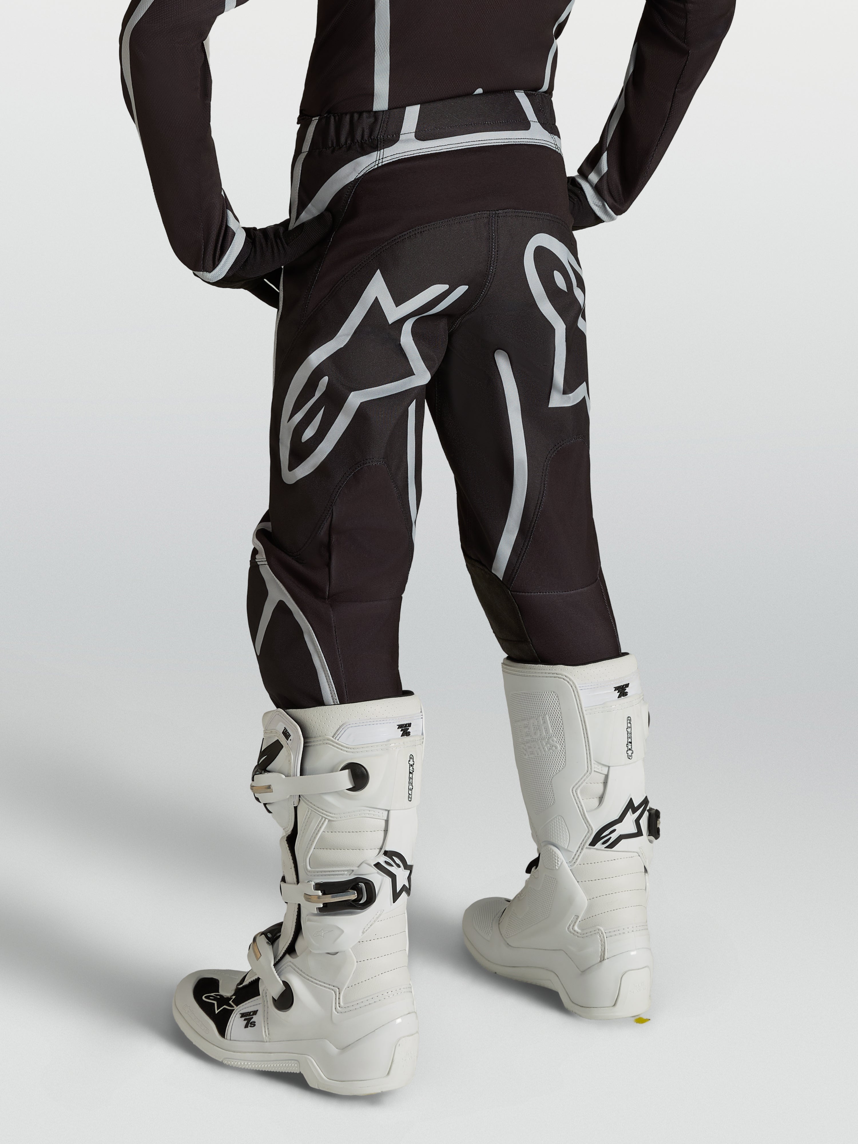 Alpinestars Calças Fluid Apex V2 Juventude, calças de motocross, pretas e prateadas, vista traseira com botas brancas.