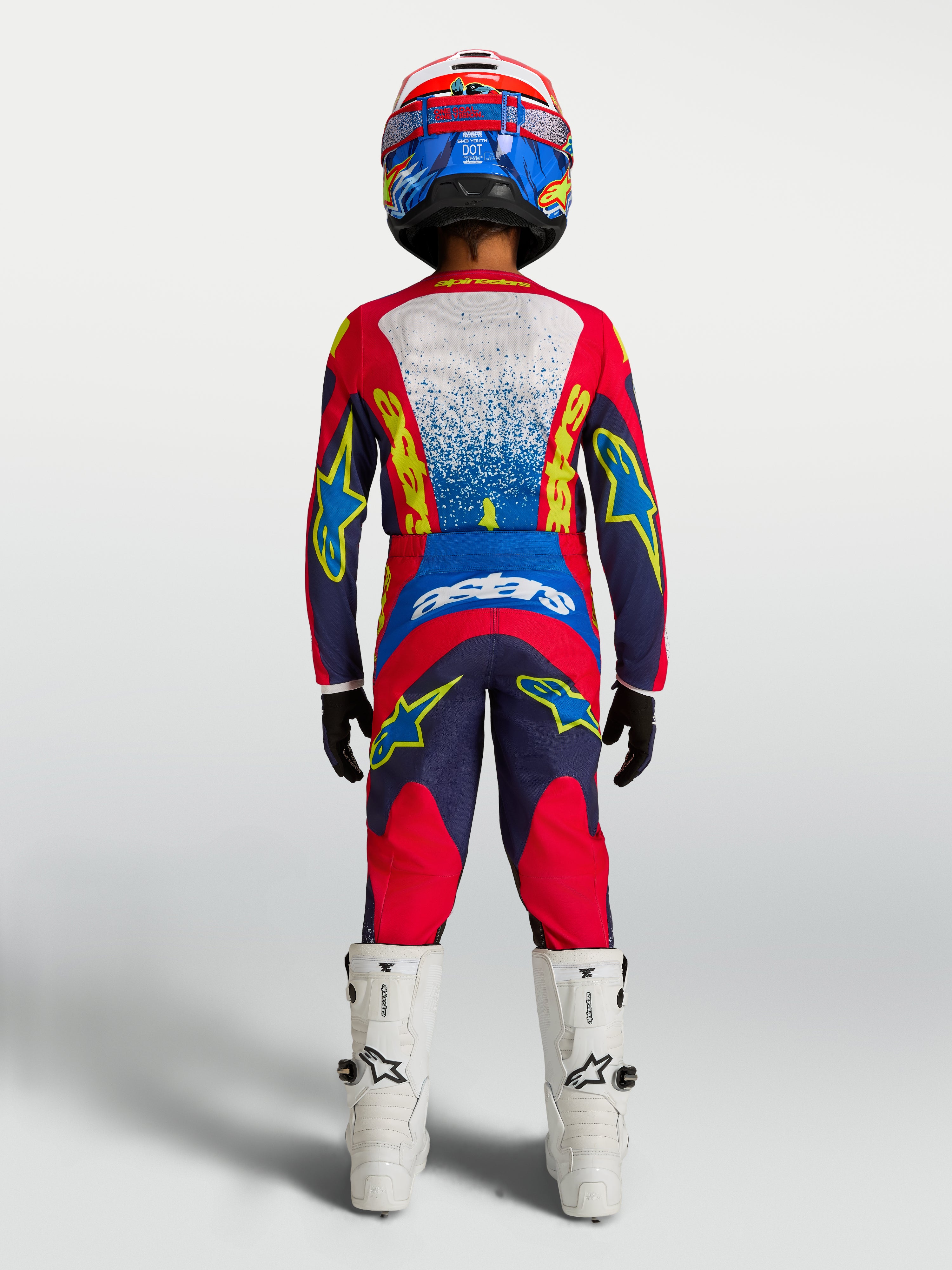 Alpinestars calças Fluid Drag juventude, equipamento de motocross azul e vermelho, vista traseira com camisola e botas brancas.