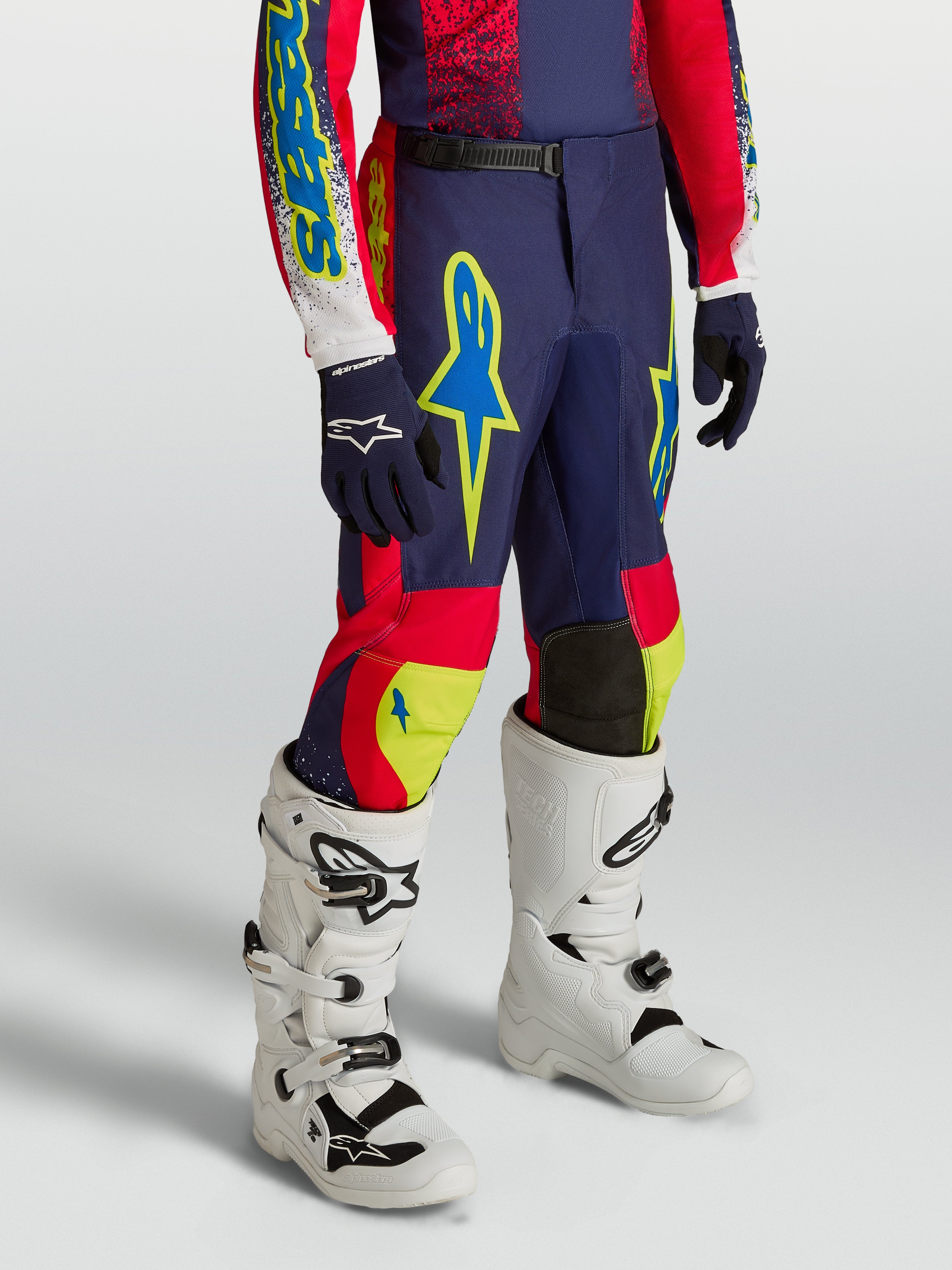 Alpinestars Calças Fluid Drag Juventude, equipamento de motocross azul-marinho e vermelho com detalhes néon e botas brancas de performance.