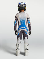 Alpinestars Calças Fluid Drag Juventude 2027, calças de motocross para jovens, cinzentas e azuis, vista traseira com conjunto de camisola e botas para condução off-road.