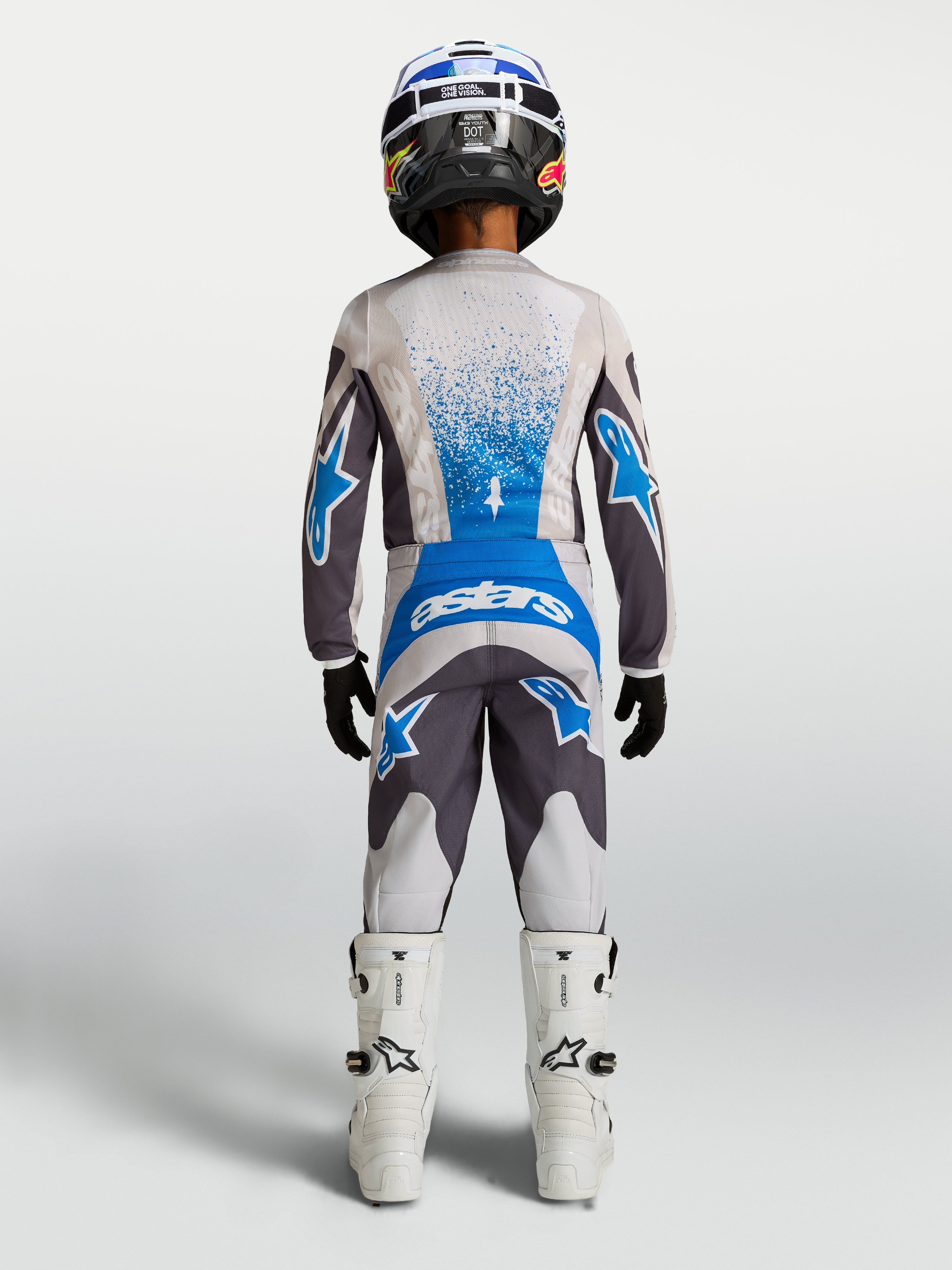 Alpinestars Calças Fluid Drag Juventude 2027, calças de motocross para jovens, cinzentas e azuis, vista traseira com conjunto de camisola e botas para condução off-road.
