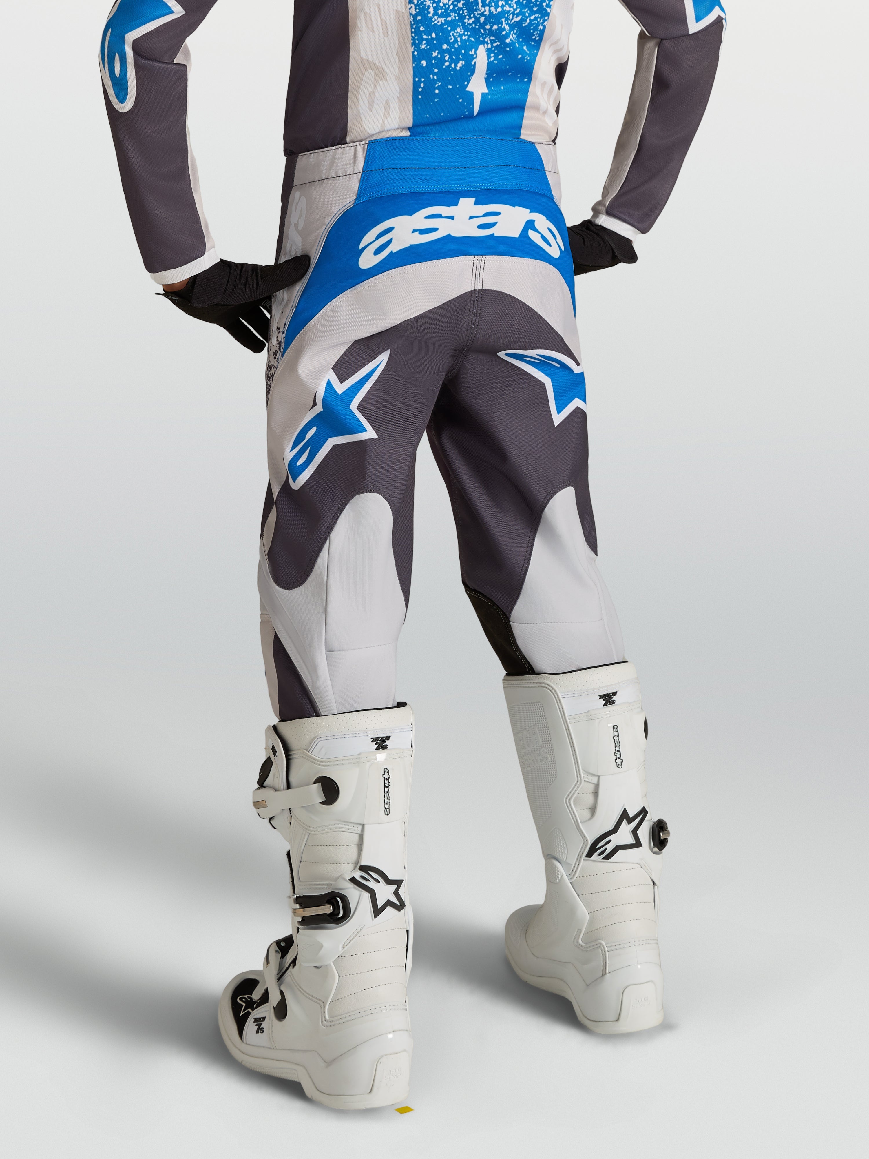 Alpinestars Calças Fluid Drag Juventude, calças de motocross cinzentas e azuis, vista traseira num piloto com botas brancas.