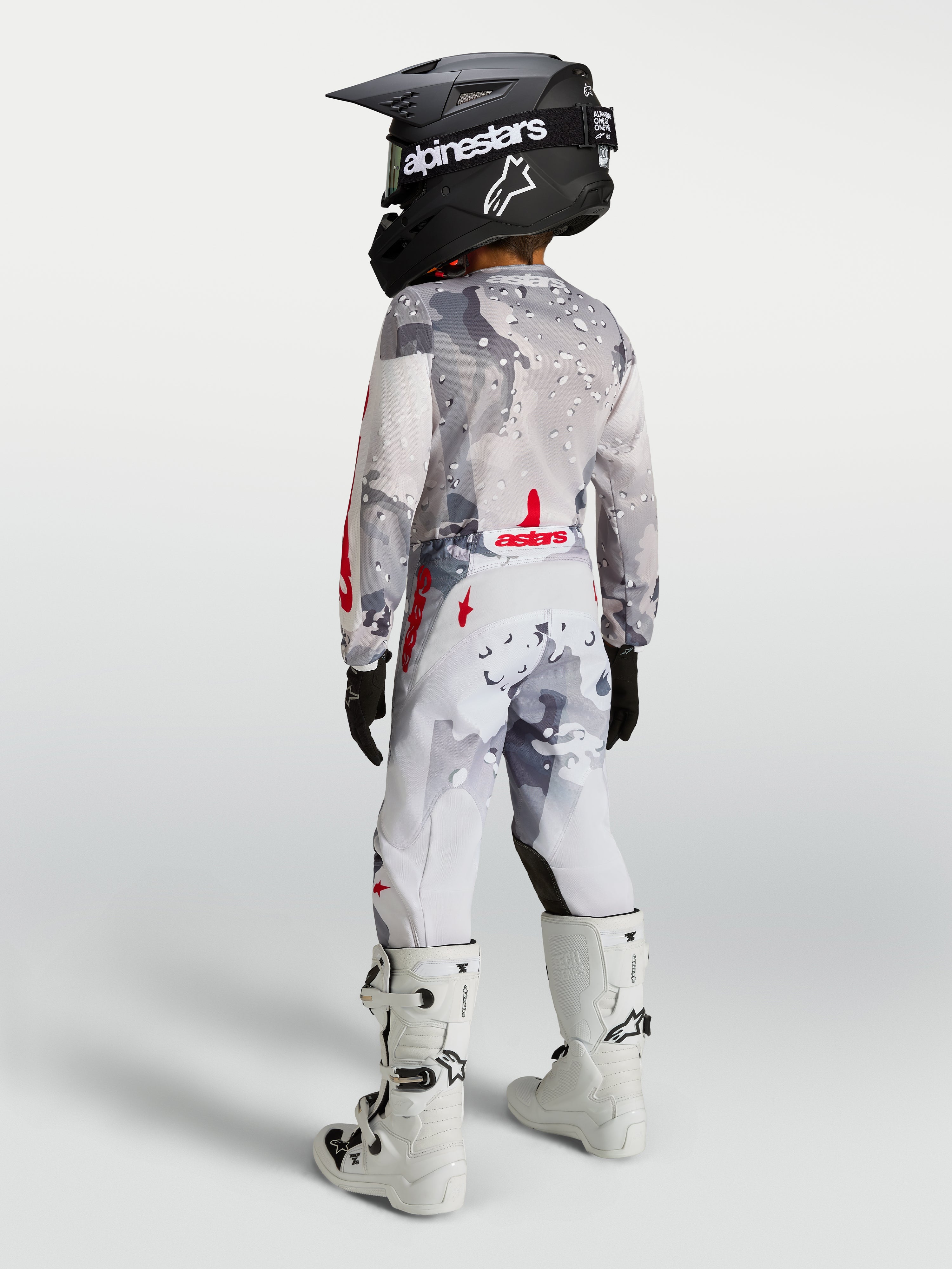 Alpinestars Calças Fluid Lone Juventude, calças de motocross e todo-o-terreno para jovens, padrão camuflagem cinzento e branco com pormenores vermelhos, vista traseira de piloto com equipamento completo.