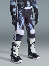 Alpinestars 2026 Youth Fluid Lahnd Pants, calças de motocross para jovens, camuflagem Iron Camo em cinzento, preto e branco, usadas com camisola de manga comprida a condizer, luvas de corrida pretas e botas de motocross brancas e pretas, com grandes logótipos de estrela nas coxas