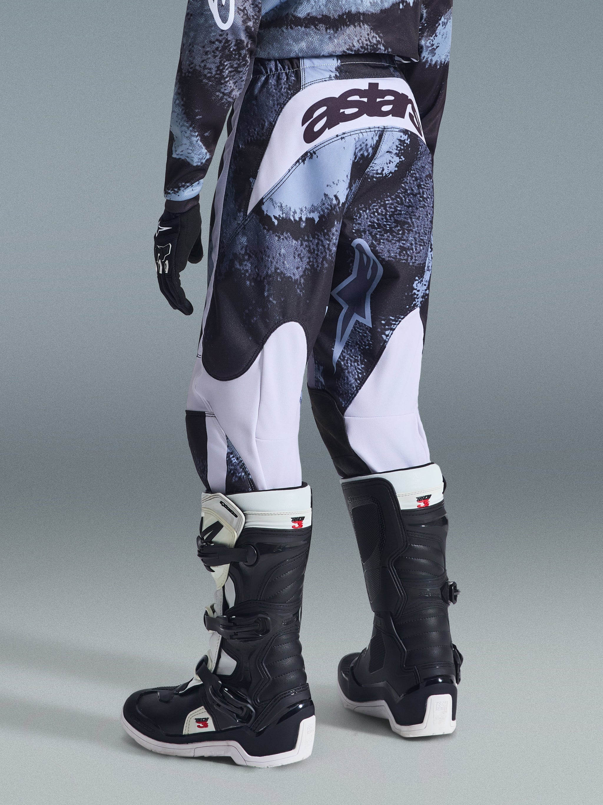 Calças Alpinestars 2026 Youth Fluid Lahnd, calças de motocross, Iron Camo com detalhes brancos, vista traseira mostrando a marca 'astars' no assento, usadas com camisola camuflada a condizer e botas de motocross Tech 3 pretas e brancas.