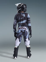 Alpinestars 2026 Youth Fluid Lahnd Pants, calças de motocross, Iron Camo com tons de azul e cinzento, vista traseira de um piloto a usar um conjunto de equipamento a condizer incluindo capacete e botas, com detalhes brancos nas pernas e logótipo 'astars' na parte de trás.