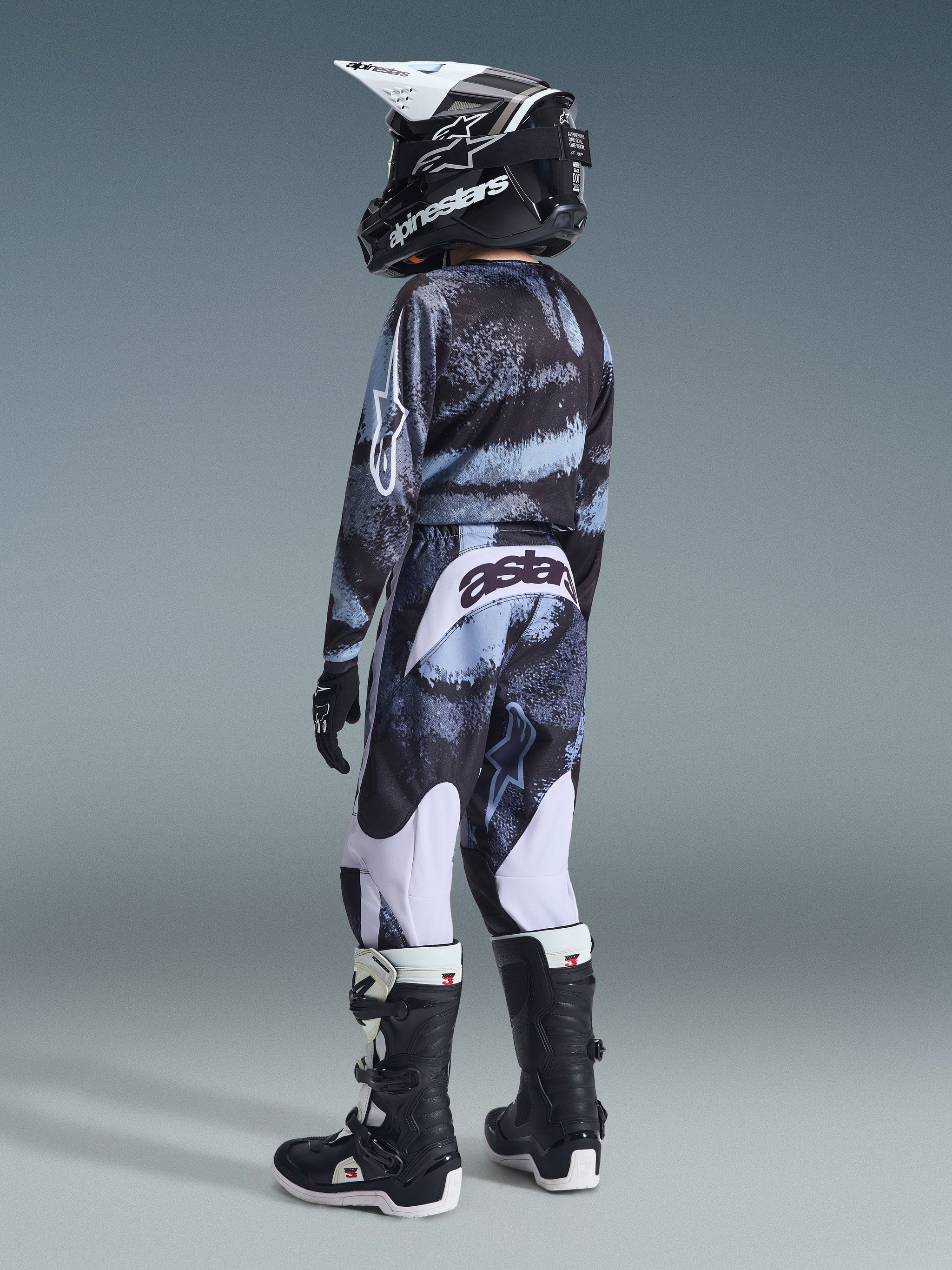 Alpinestars 2026 Youth Fluid Lahnd Pants, calças de motocross, padrão Iron Camo, piloto jovem visto de costas com camisola camuflada a condizer, botas pretas e brancas e capacete preto, com painéis brancos e marca 'astars' na parte de trás, conjunto completo de equipamento para corridas off-road