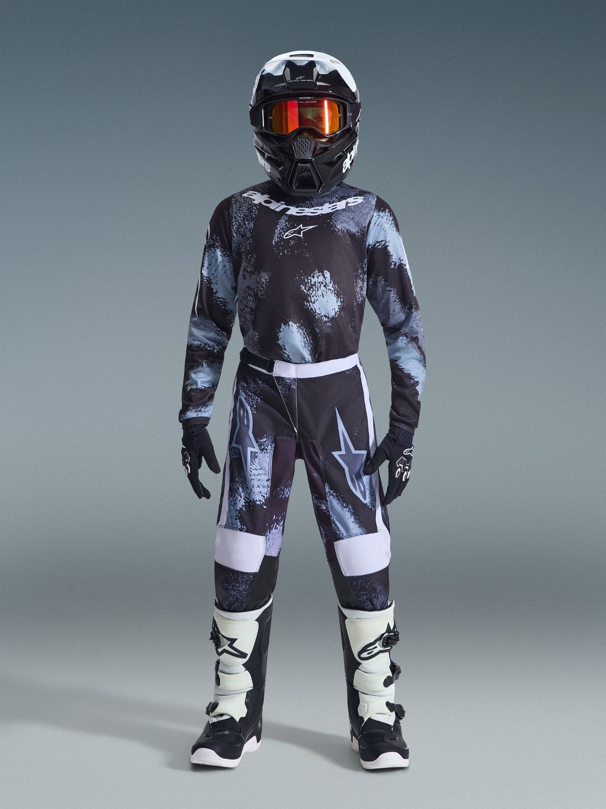 Alpinestars 2026 Youth Fluid Lahnd Pants, calças de motocross, Iron Camo, usadas por um jovem piloto com camisola de camuflagem a condizer, capacete preto com óculos cor de laranja e botas brancas e pretas, com um design coordenado, logótipos marcantes e painéis reforçados para competição fora de estrada